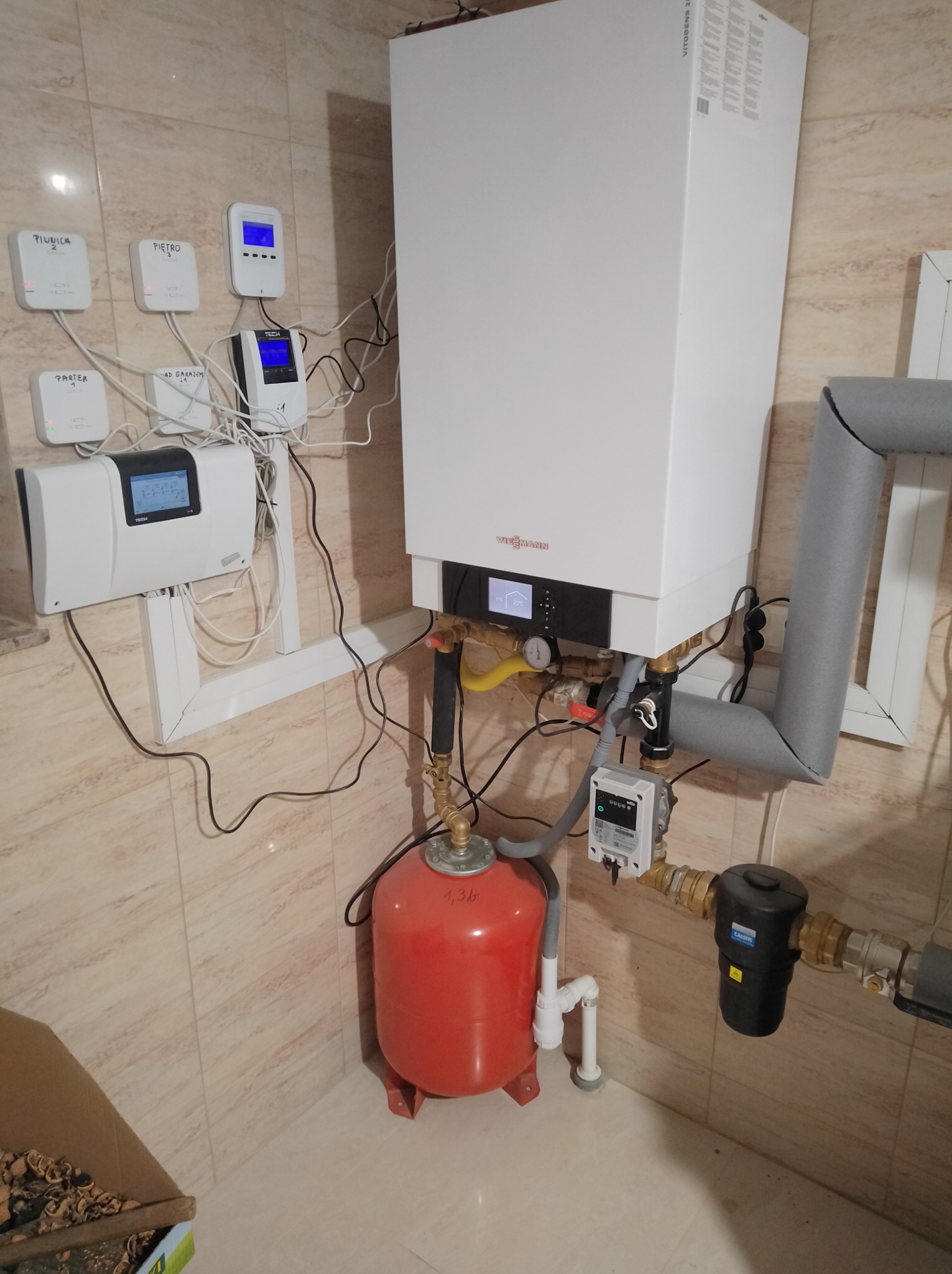 Biały piec gazowy Viessmann zamontowany na ścianie z beżowych płytek, z widocznymi przewodami, czerwonym zbiornikiem i panelami sterowania. Widok z dołu.