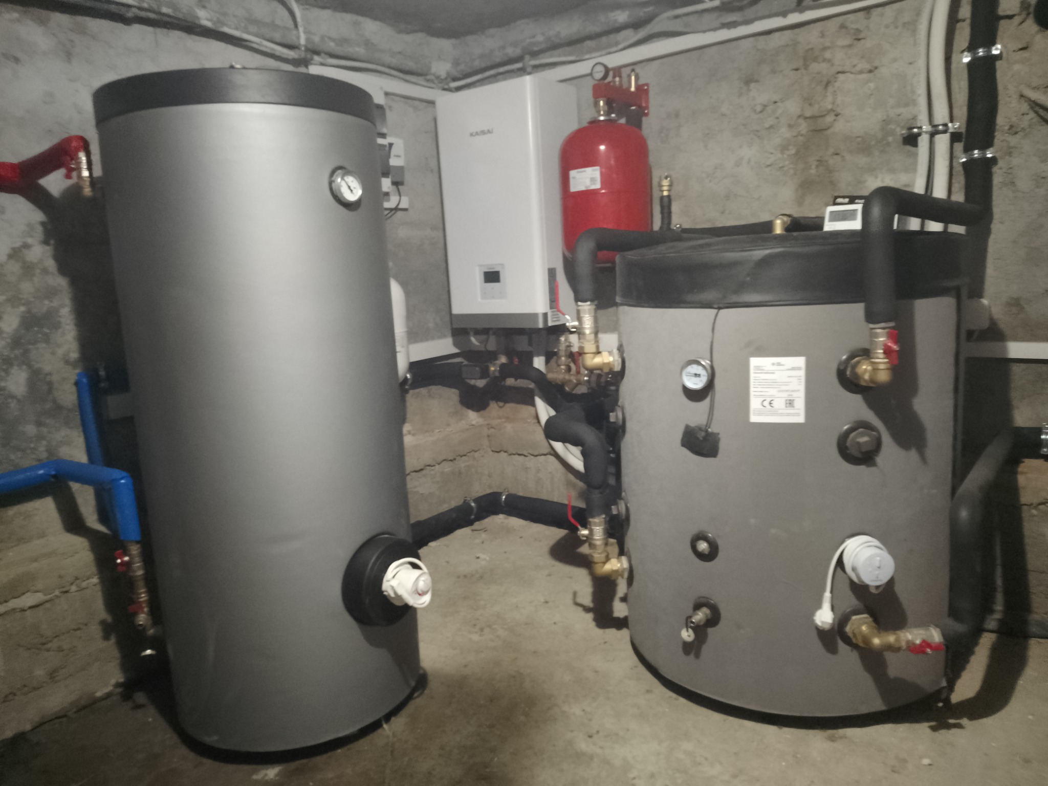 Dwa zbiorniki akumulacyjne w szarej obudowie, z widocznymi przyłączami hydraulicznymi i elementami instalacji grzewczej, zainstalowane w pomieszczeniu z betonowymi ścianami.