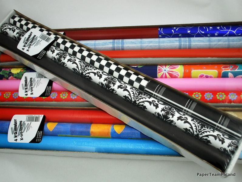 Zestaw ozdobnych papierów w rolkach, widoczne wzory: szachownica i motywy roślinne, zapakowane w podłużne pudełko, ułożone warstwami z innymi kolorowymi papierami.