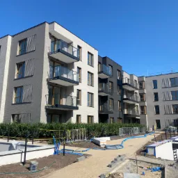 Nowoczesny budynek wielorodzinny z elewacją z szarej, elastycznej płytki klinkierowej Elastolith, balkony z szklanymi balustradami, widoczne elementy placu budowy i niebieskie węże doprowadzające...