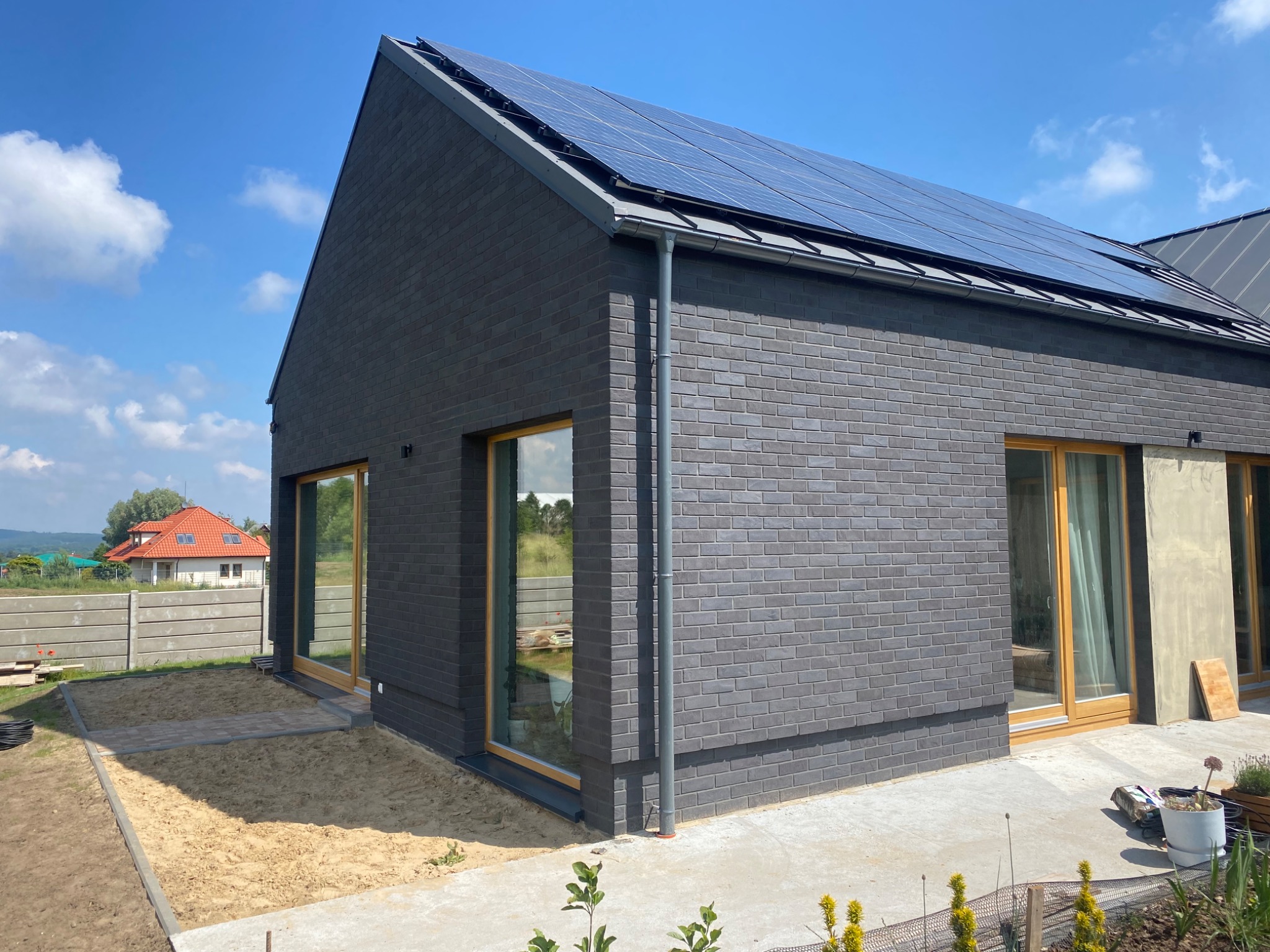 Nowoczesny dom jednorodzinny z ciemnej cegły klinkierowej z panelami słonecznymi na dachu, widok z zewnątrz na tle błękitnego nieba.