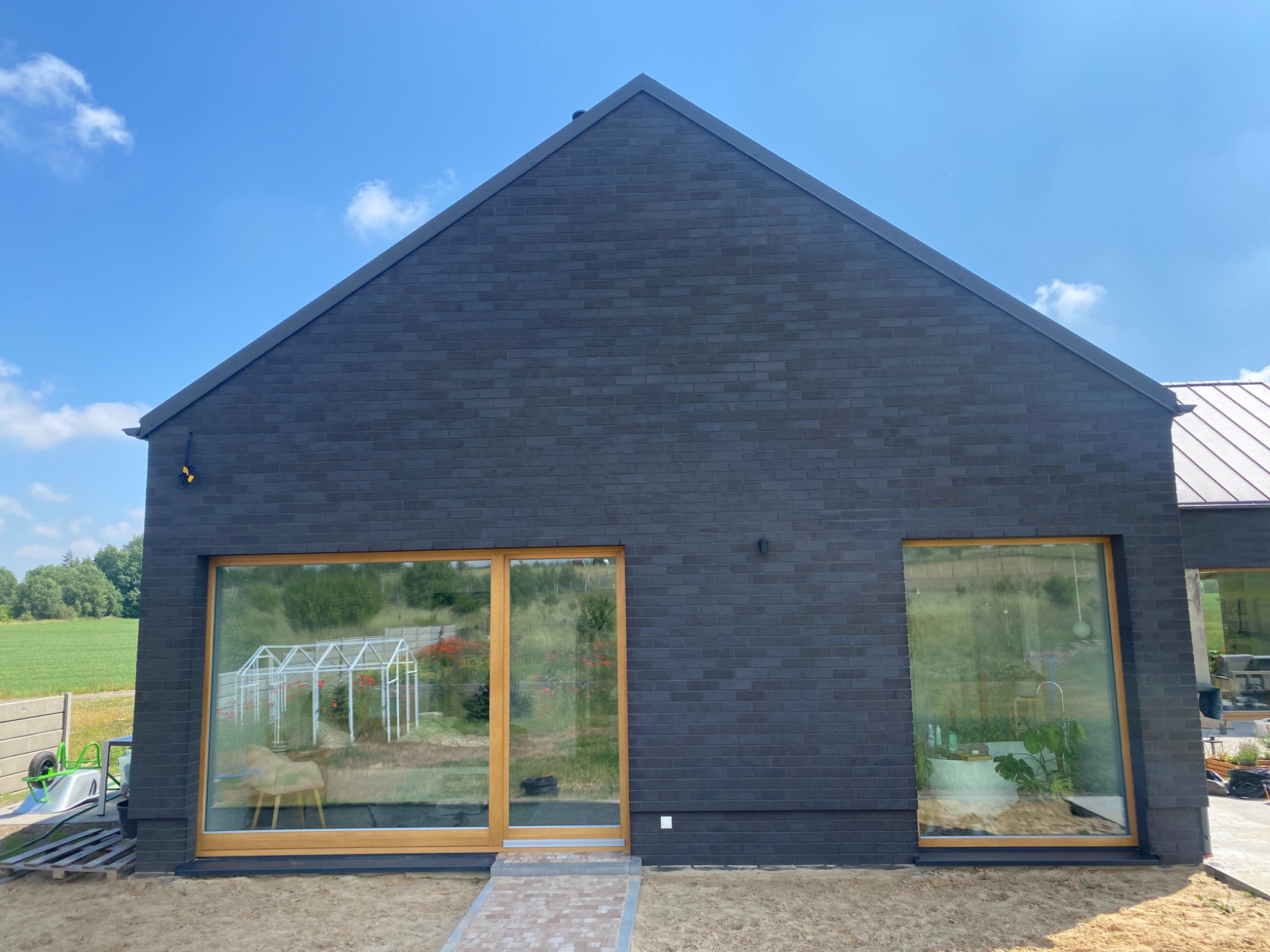 Nowoczesny dom jednorodzinny z ciemnej cegły klinkierowej, duże okna z drewnianą ramą, widok na ogród i szklarnię, ścieżka z kostki brukowej.