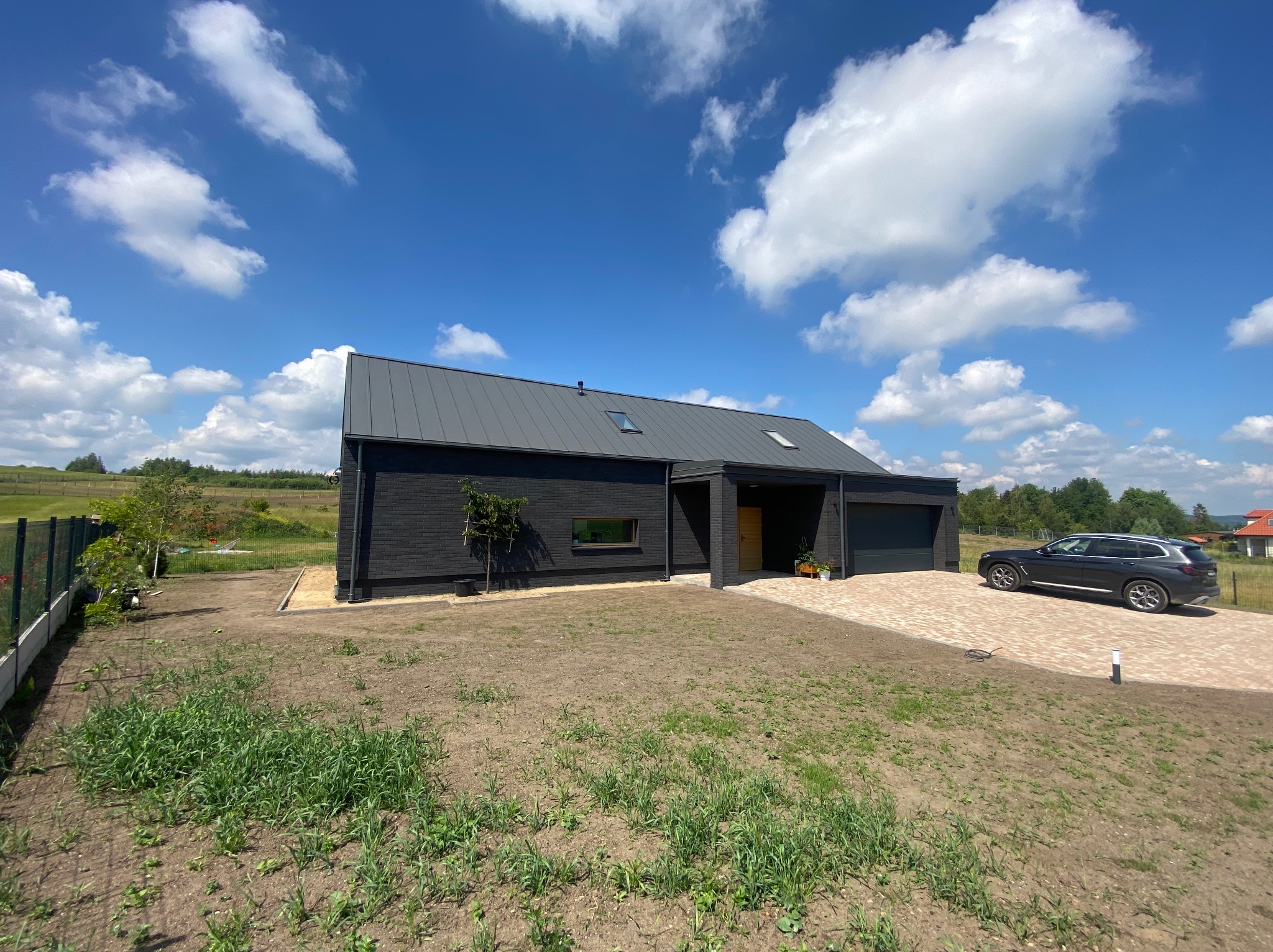 Nowoczesny dom jednorodzinny z ciemną elewacją, szarym dachem i brukowanym podjazdem w słoneczny dzień z błękitnym niebem i białymi chmurami.