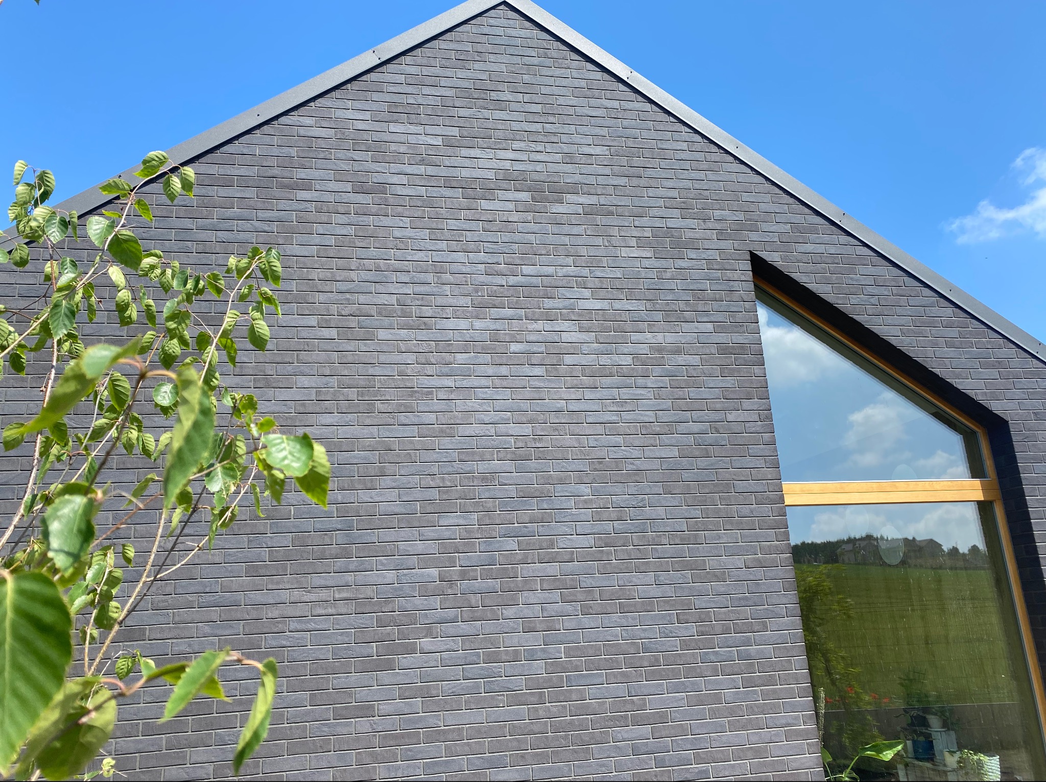 Nowoczesny dom jednorodzinny z elewacją z ciemnoszarej, elastycznej płytki klinkierowej ELASTOLITH, widoczne asymetryczne okno z drewnianą ramą i fragment drzewa z zielonymi liśćmi na tle...