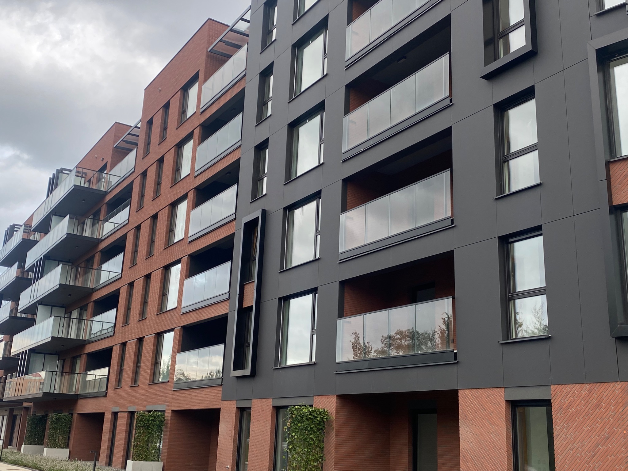 Nowoczesny budynek wielorodzinny z elewacją z klinkieru i paneli w kolorze antracytowym, z balkonami z szklanymi balustradami i zielenią u podstawy.