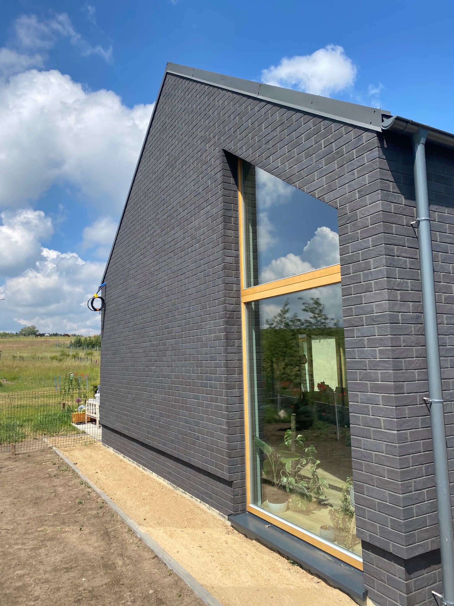 Nowoczesna elewacja domu wykończona ciemną, elastyczną płytką klinkierową ELASTOLITH, z widocznym narożnikiem, oknem z drewnianą ramą i rynną w kolorze grafitowym oraz fragmentem trawnika w tle.