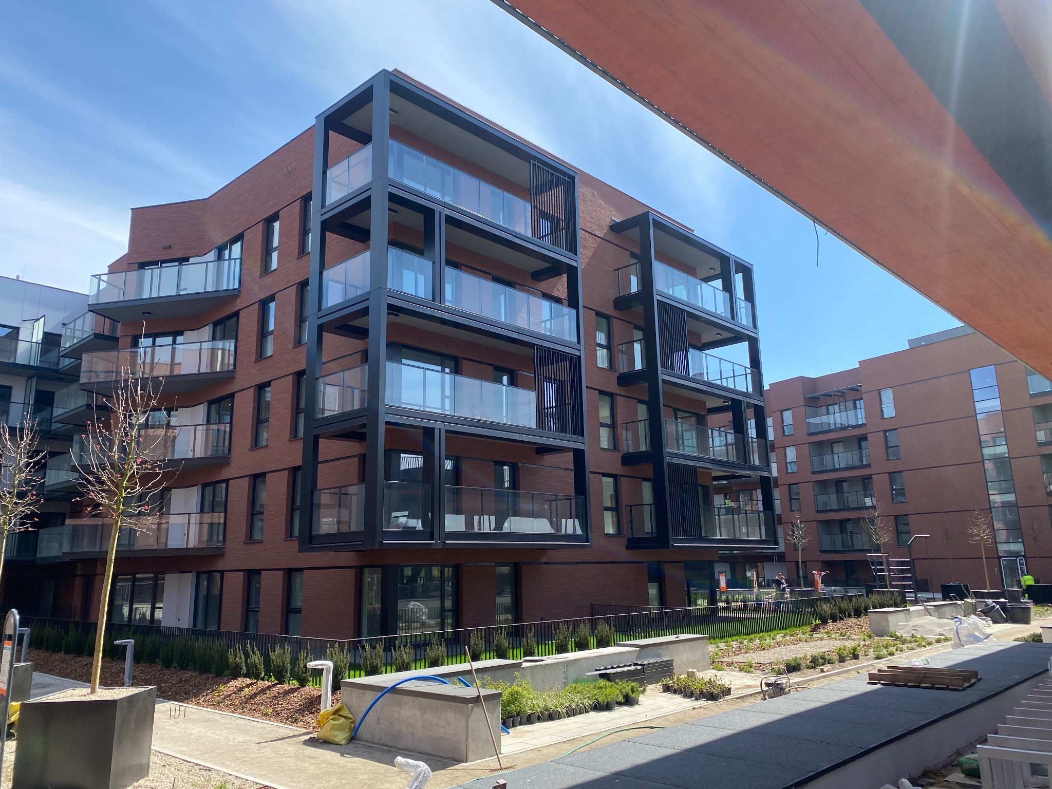 Nowoczesny budynek mieszkalny z elewacją z czerwonej cegły klinkierowej i czarnymi ramami balkonów, widok z poziomu gruntu na plac budowy z elementami małej architektury i roślinnością w trakcie...