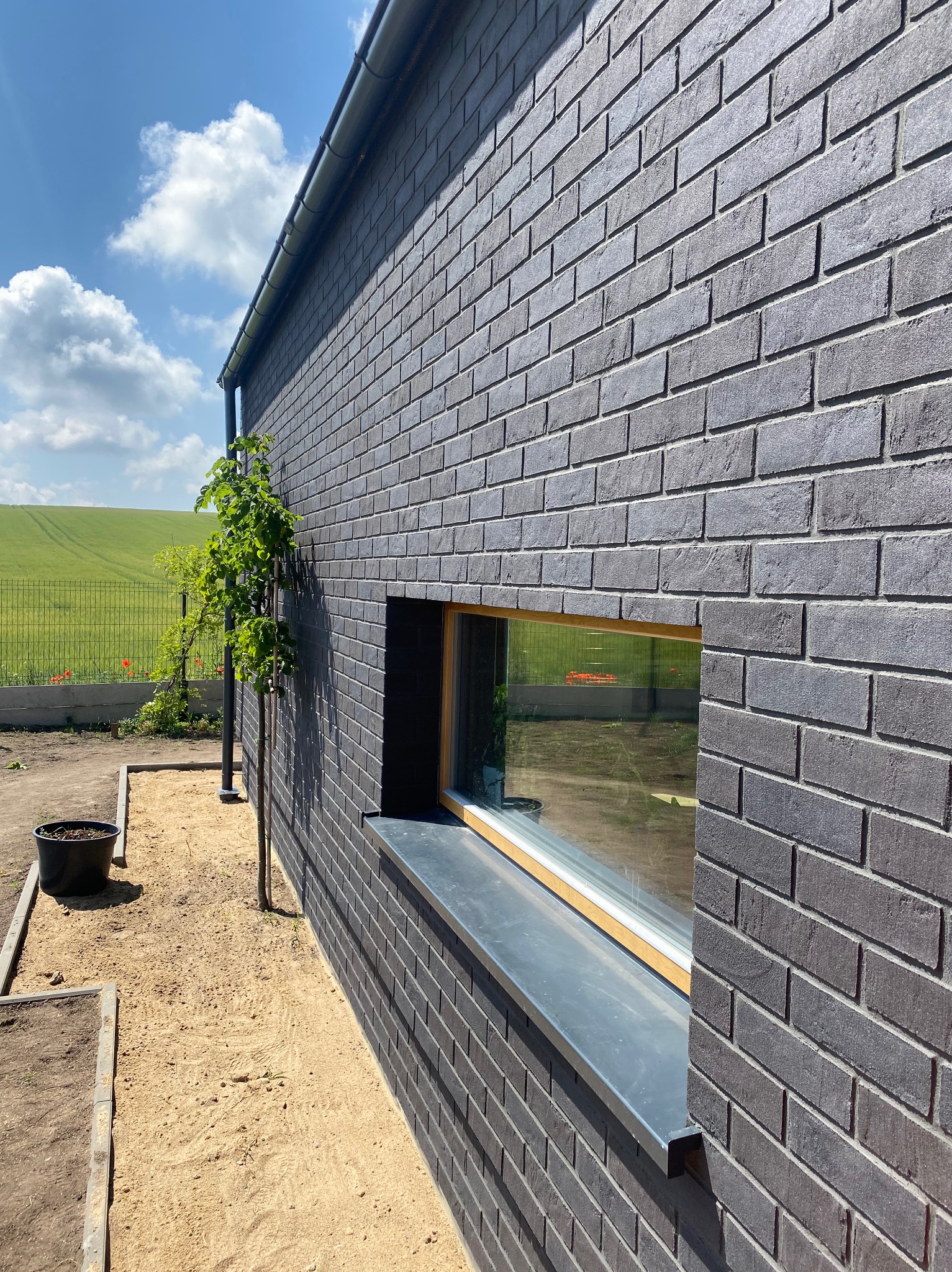 Szara elewacja z cegły klinkierowej z oknem, widok na zielone pole i błękitne niebo, małe drzewko przy ścianie, piasek przy elewacji.