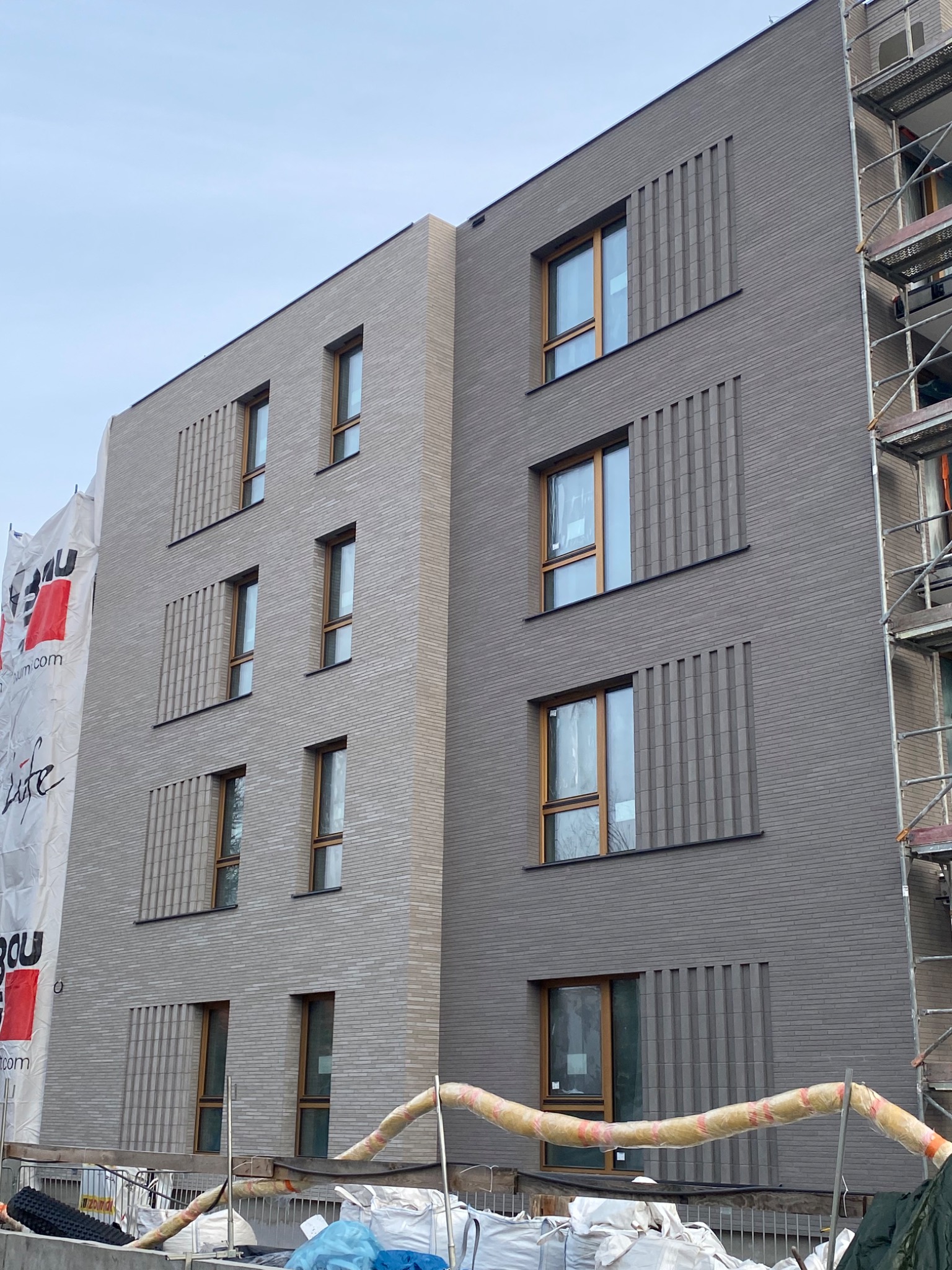 Nowoczesna elewacja budynku wielorodzinnego z szarej, elastycznej płytki klinkierowej, z widocznymi oknami w drewnianych ramach i elementami dekoracyjnymi w postaci pionowych pasków. Na pierwszym...