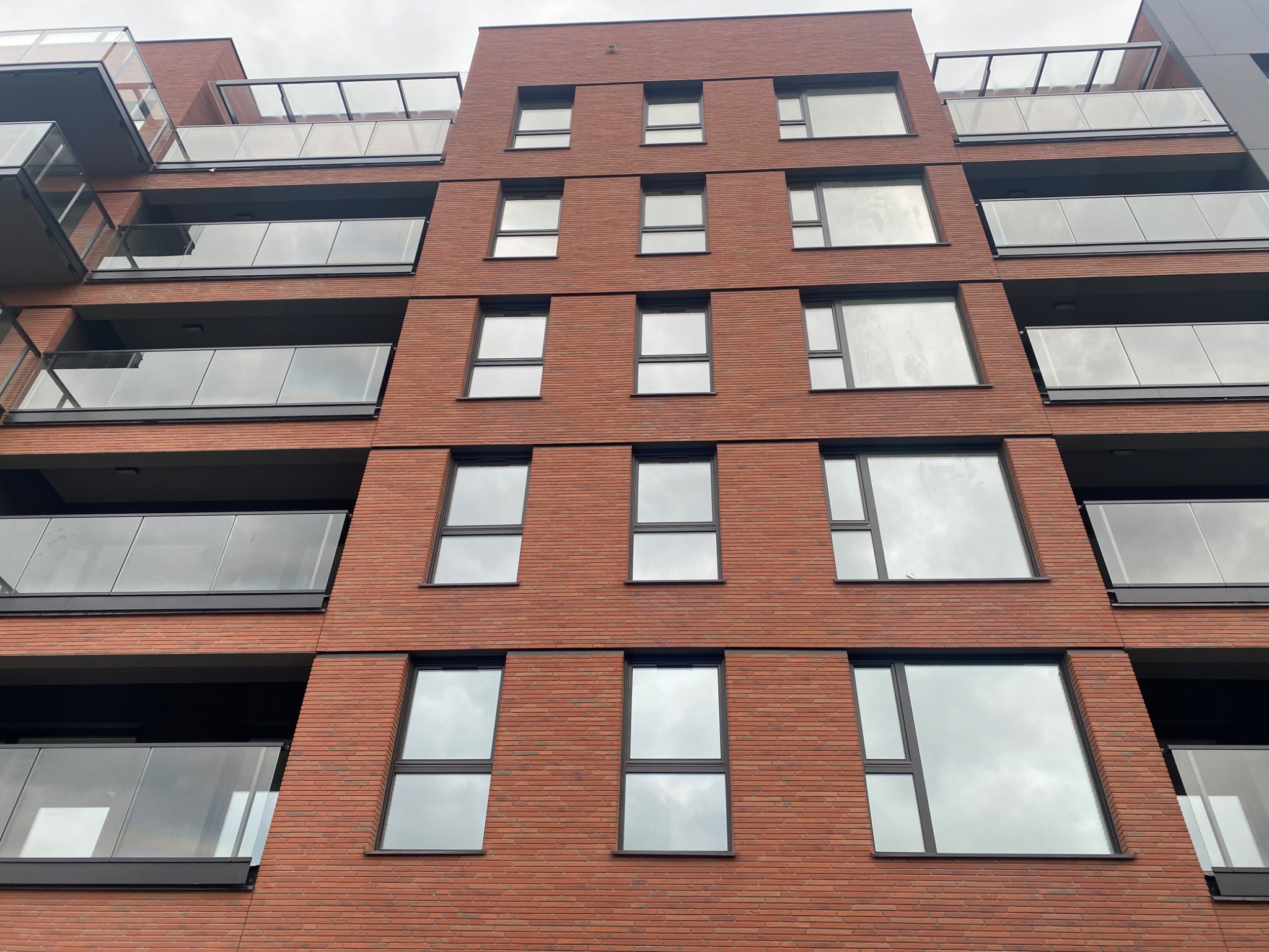Fasada budynku wielorodzinnego wykończona czerwoną, elastyczną płytką klinkierową imitującą cegłę, z balkonami z szklanymi balustradami i oknami w ciemnych ramach.