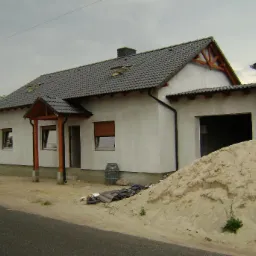 Dom jednorodzinny z ciemną dachówką ceramiczną, widoczne rynny, więźba dachowa i sterta piasku przed budynkiem, częściowo wykończona elewacja