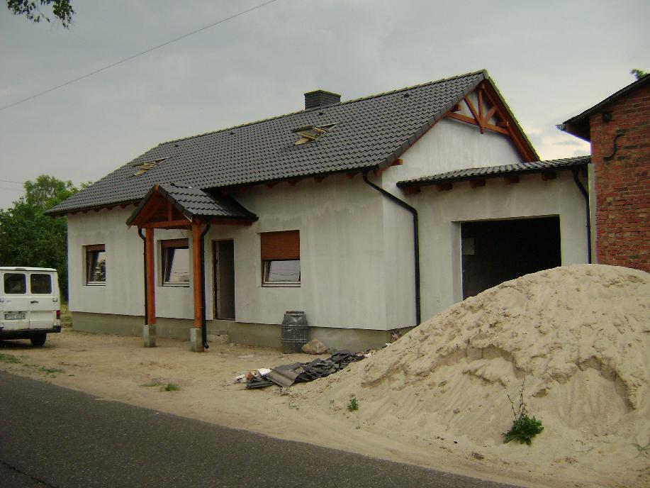 Dom jednorodzinny z ciemną dachówką ceramiczną, widoczne rynny, więźba dachowa i sterta piasku przed budynkiem, częściowo wykończona elewacja