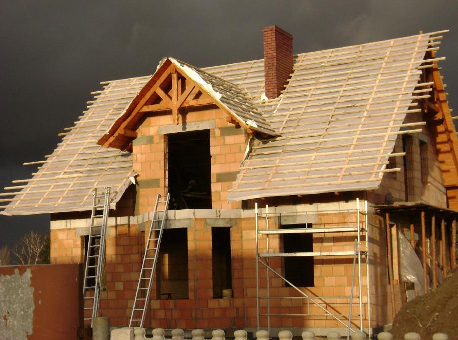 Dom w trakcie budowy z widoczną konstrukcją więźby dachowej pod folią, ceglany komin, rusztowania i drabiny, na tle ciemnego nieba.
