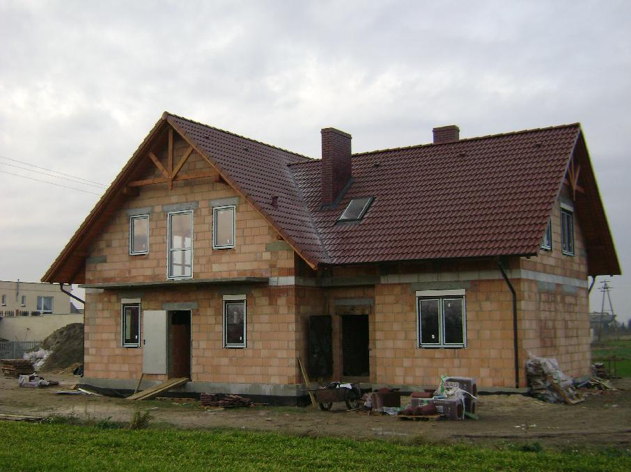 Dom w trakcie budowy z widoczną konstrukcją dachu z drewnianą więźbą i częściowo położoną czerwoną dachówką, komin z cegły, okno dachowe oraz nieotynkowane ściany z pustaków ceramicznych.