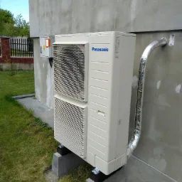 Zewnętrzna jednostka klimatyzacji Panasonic zamontowana na betonowych podstawach przy szarej ścianie budynku z widocznymi przewodami w izolacji aluminiowej.