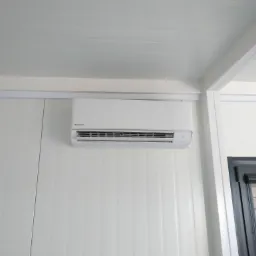 Biała jednostka klimatyzacyjna Panasonic zamontowana na białej, panelowej ścianie wewnętrznej, widoczna część okna z żaluzjami.