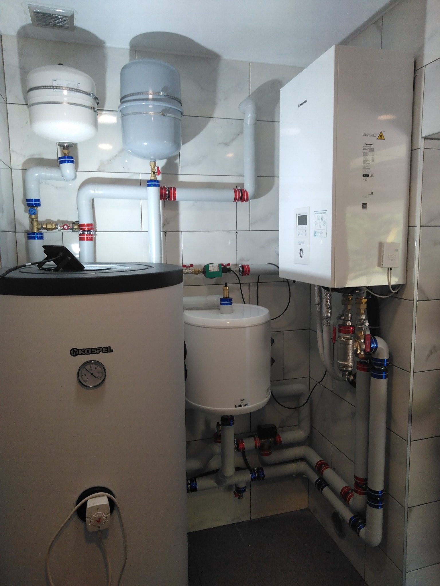 Zainstalowany system grzewczy w kotłowni z białymi rurami z niebieskimi i czerwonymi złączkami, zbiornikiem Kospel z manometrem, białym piecem Panasonic i dwoma zbiornikami wyrównawczymi na tle...
