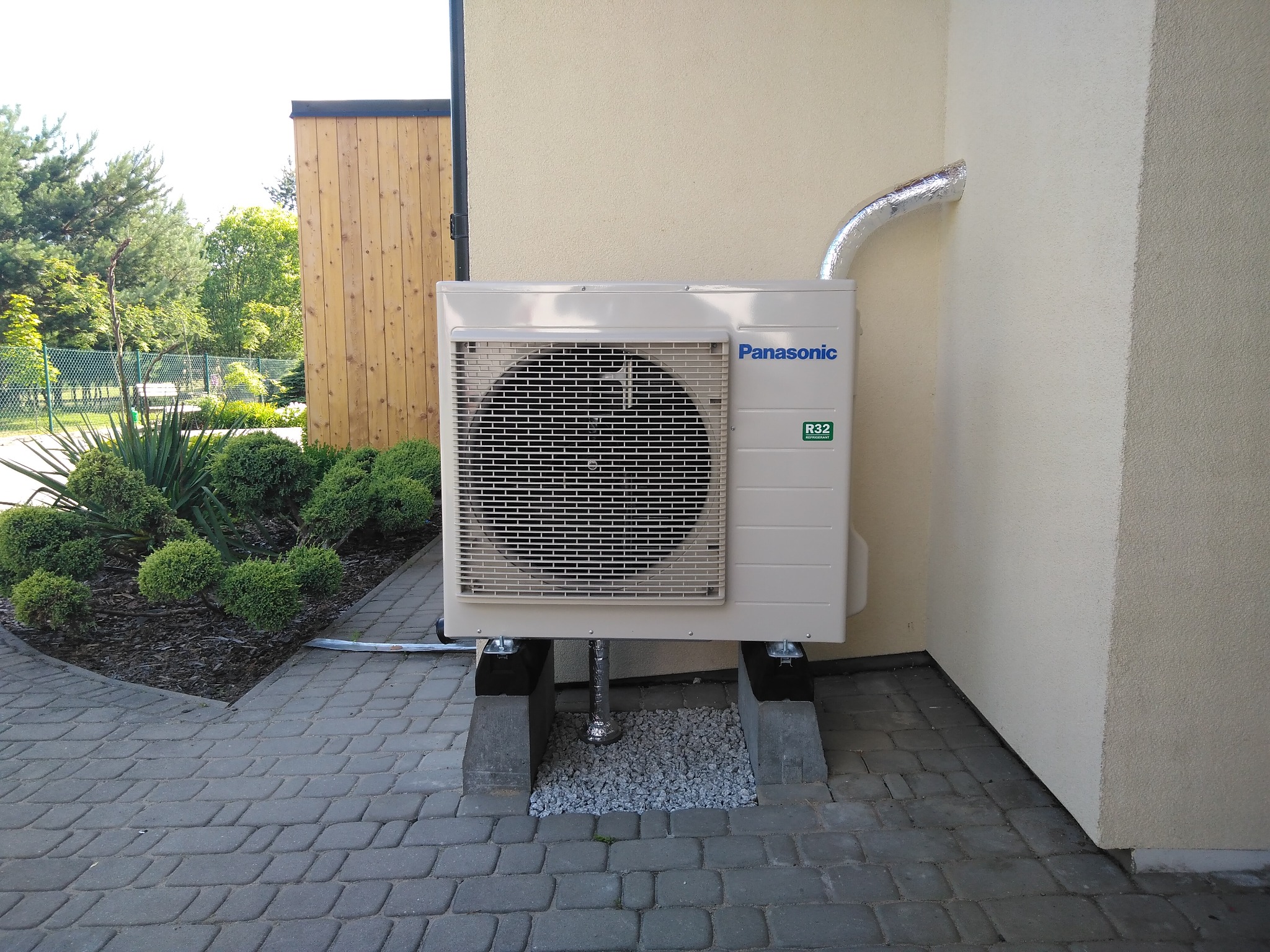 Zewnętrzna jednostka klimatyzacji Panasonic z widocznym logo i oznaczeniem R32, zamontowana na betonowych podstawach z podsypką żwirową, na tle elewacji budynku i kostki brukowej.