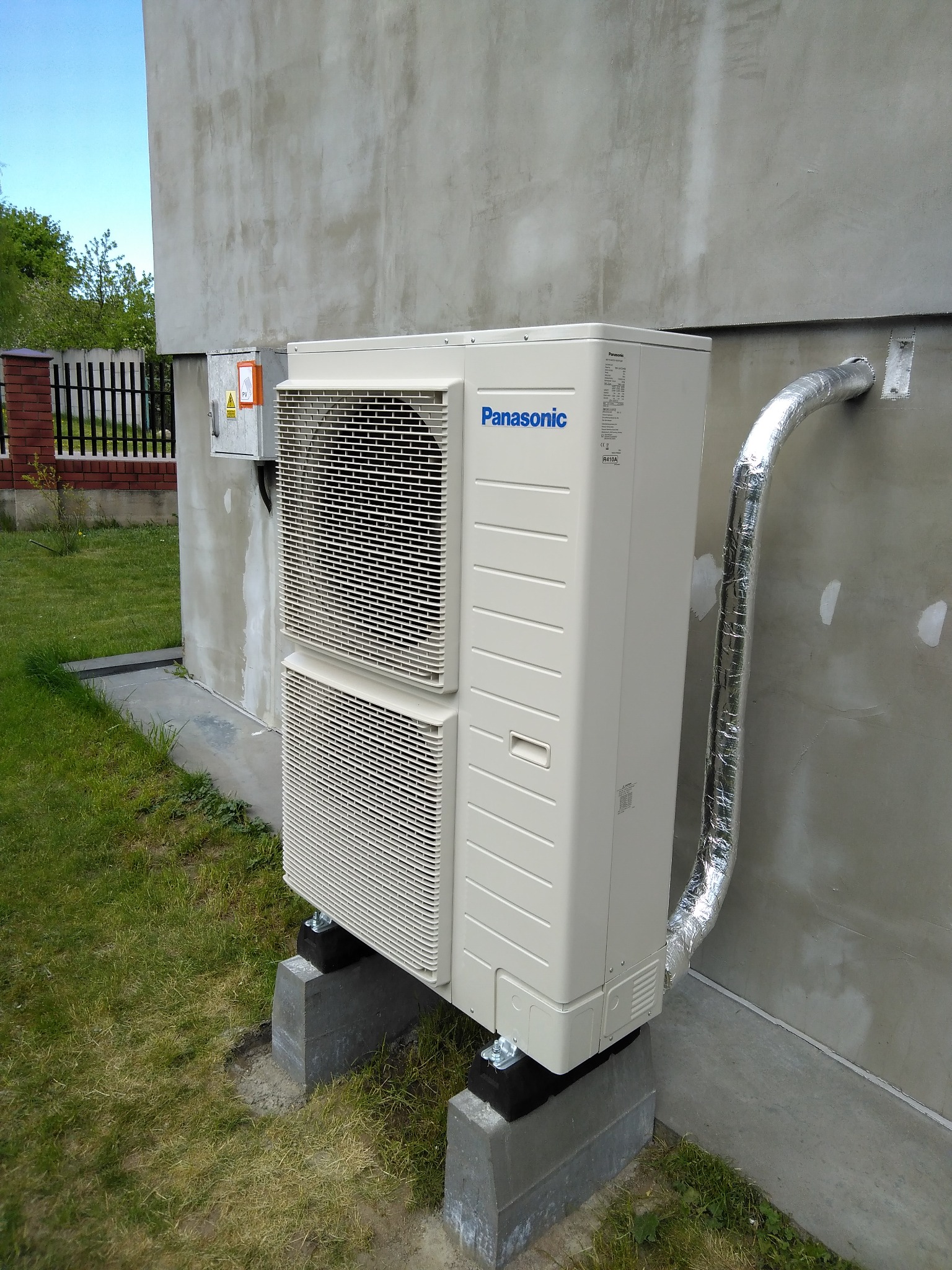 Zewnętrzna jednostka klimatyzacji Panasonic zamontowana na betonowych podstawach przy szarej ścianie budynku z widocznymi przewodami w izolacji aluminiowej.
