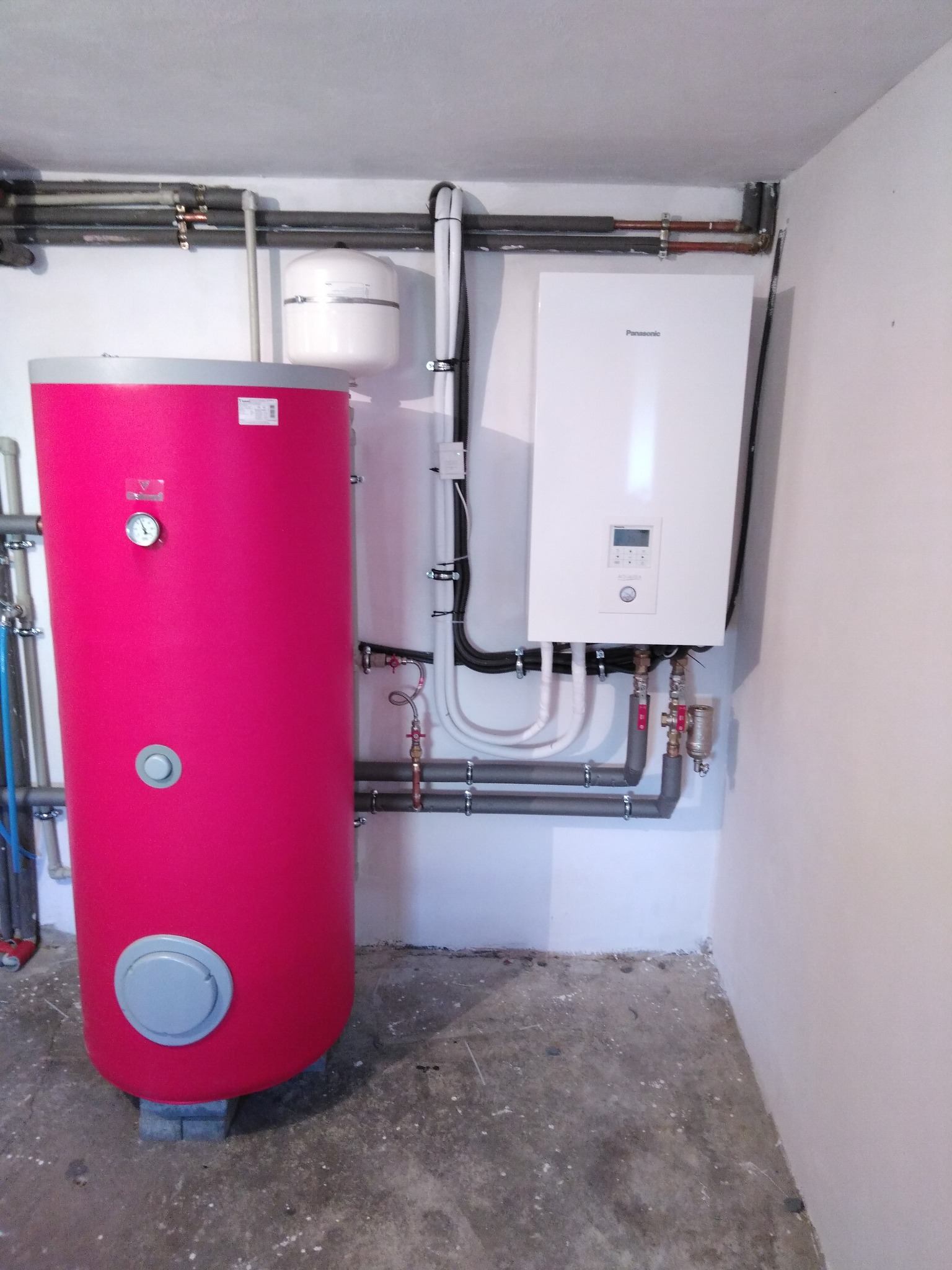 Instalacja grzewcza w piwnicy: Czerwony zasobnik CWU z manometrem obok białego pieca gazowego Panasonic, rury miedziane i plastikowe prowadzone wzdłuż ścian, surowy beton na podłodze.