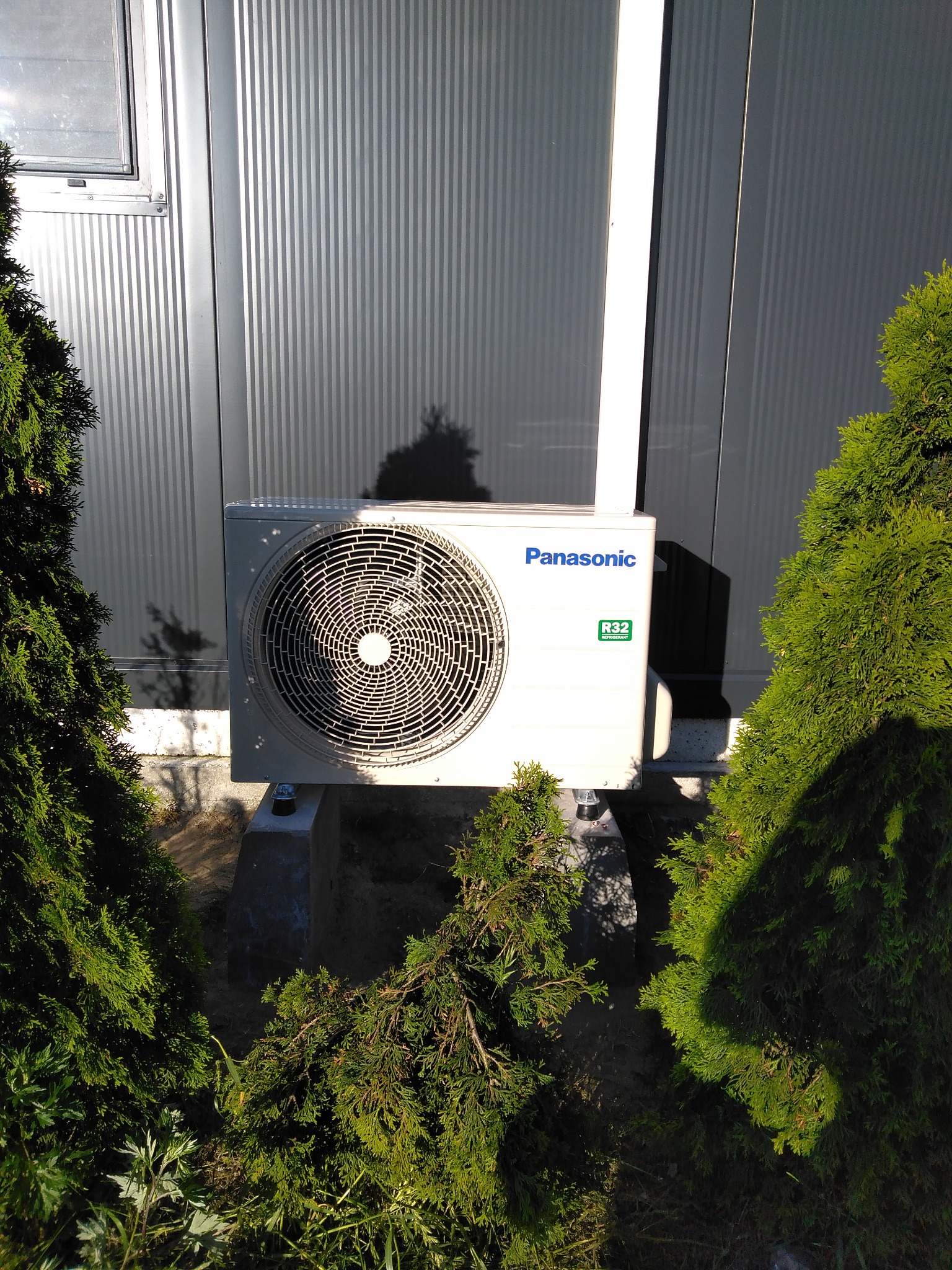 Zewnętrzna jednostka klimatyzacji Panasonic R32 zamontowana na betonowych podstawach, otoczona tujami, przy metalowej ścianie budynku.