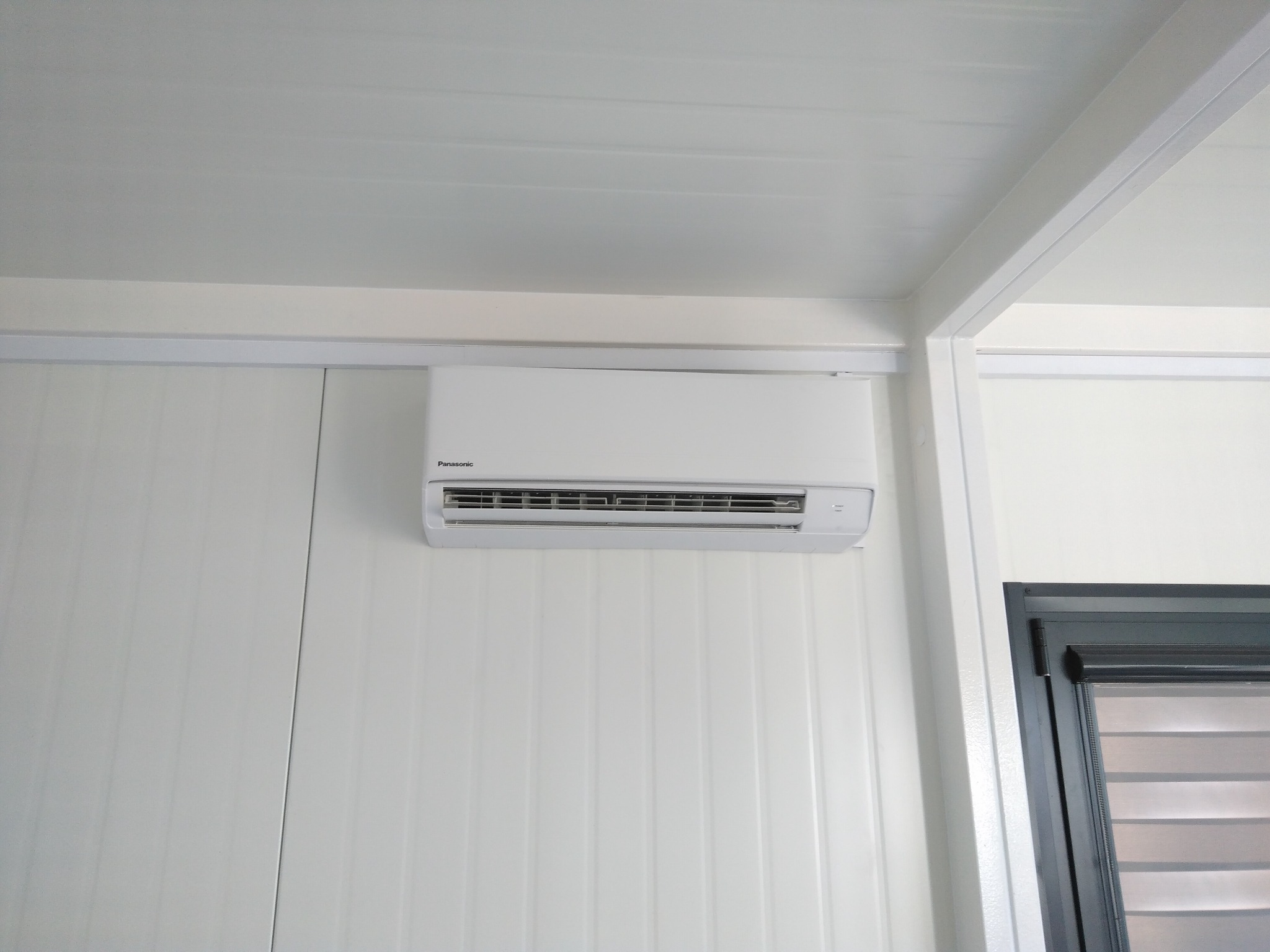 Biała jednostka klimatyzacyjna Panasonic zamontowana na białej, panelowej ścianie wewnętrznej, widoczna część okna z żaluzjami.