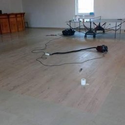 Remonty.ii.wykonczenia - Fragment podłogi z paneli w trakcie układania, widoczny odkurzacz, rolka taśmy malarskiej i stół do ping-ponga w tle.