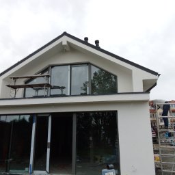 Remonty.ii.wykonczenia - Nowoczesny dom w trakcie wykończenia elewacji, z widocznym rusztowaniem i pracownikiem na drabinie, duże okna od podłogi do sufitu, szare niebo w tle.