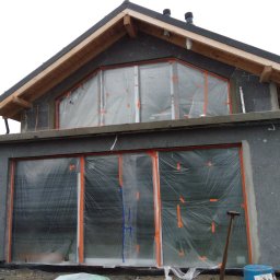 Remonty.ii.wykonczenia - Surowa elewacja budynku w trakcie ocieplania, okna zabezpieczone folią, widoczne narzędzia i materiały budowlane.