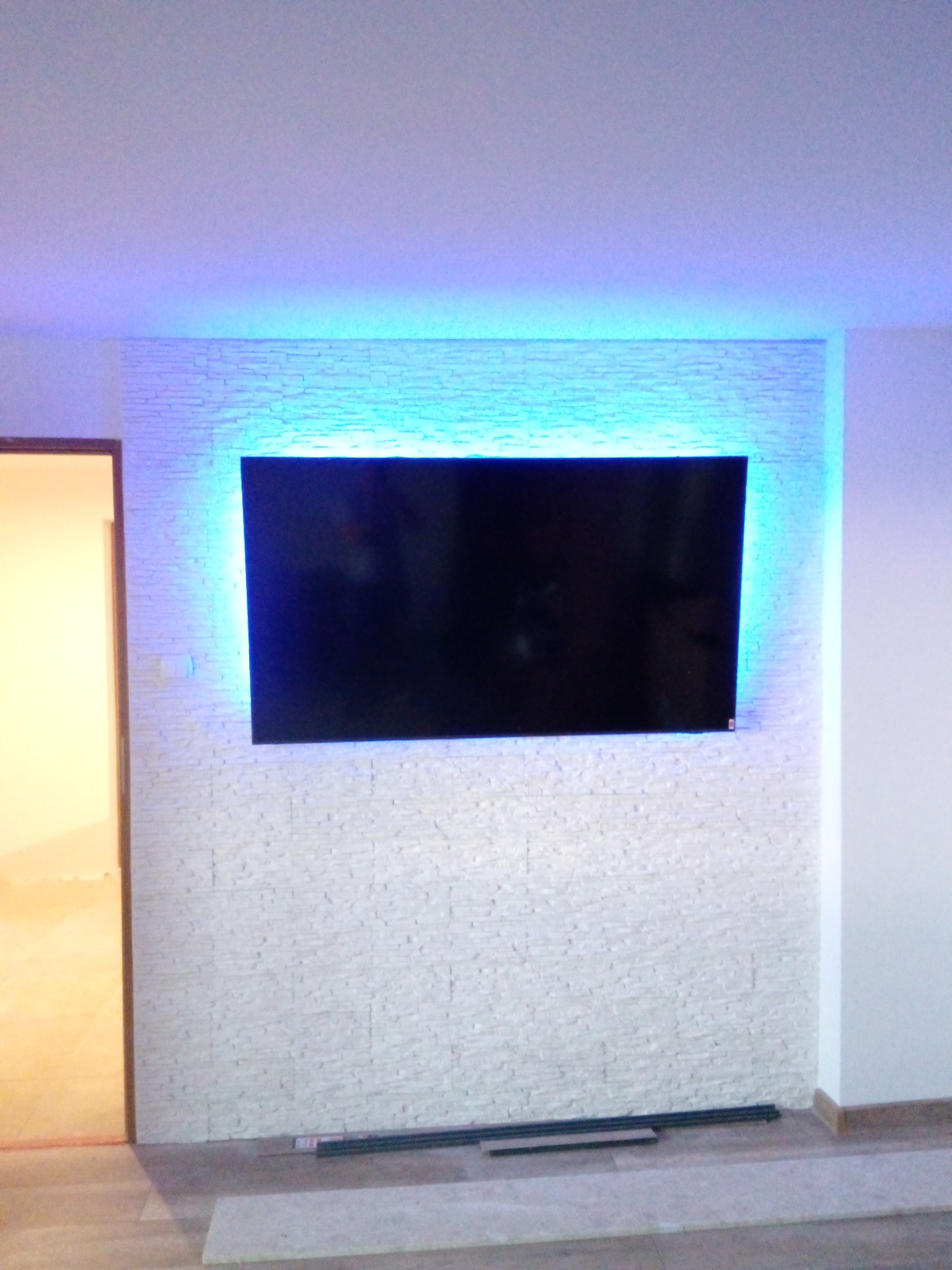 Telewizor zawieszony na ścianie wyłożonej dekoracyjną, białą cegłą z niebieskim podświetleniem LED, widoczne listwy przypodłogowe na podłodze.