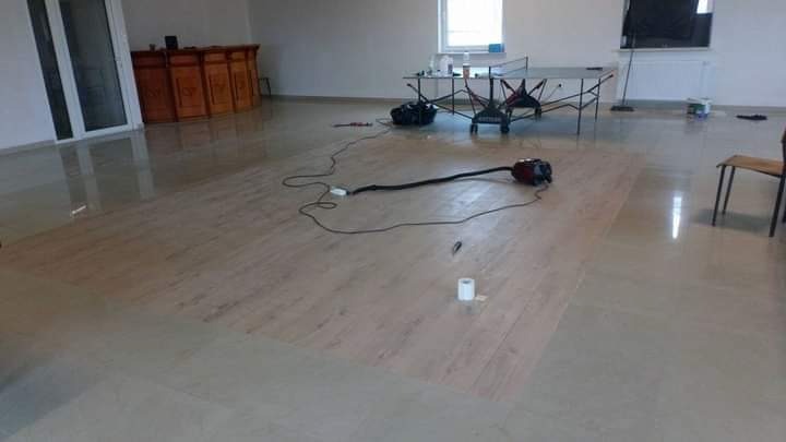 Fragment podłogi z paneli w trakcie układania, widoczny odkurzacz, rolka taśmy malarskiej i stół do ping-ponga w tle.