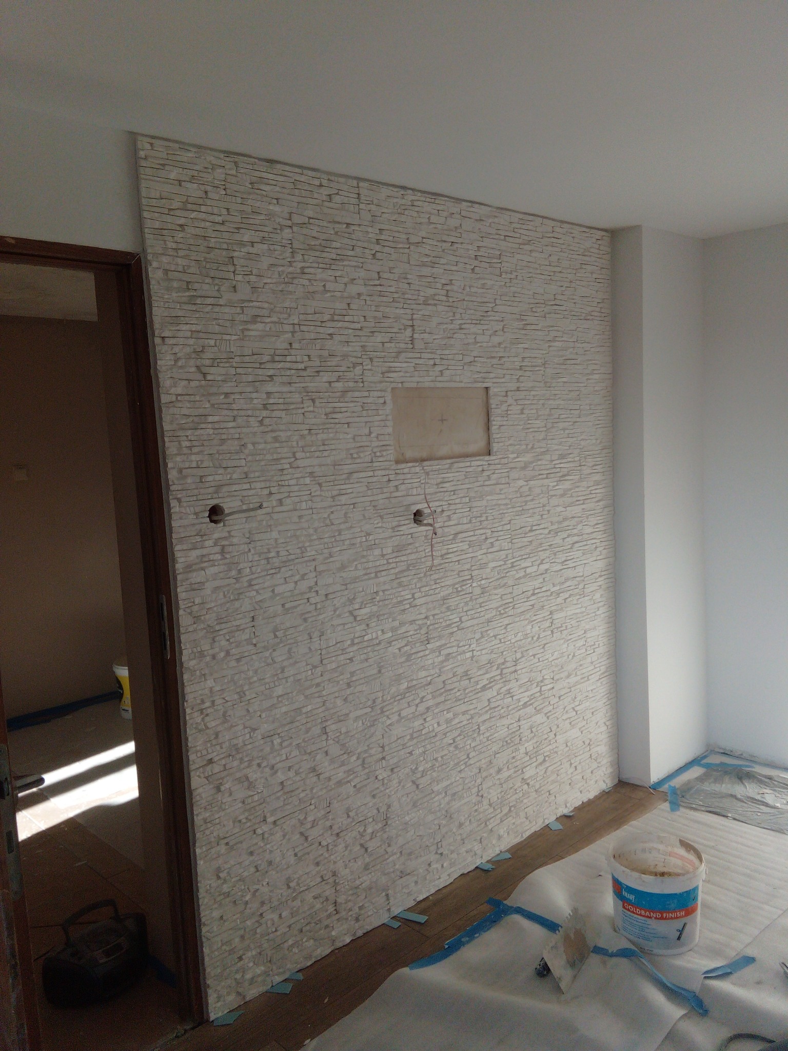 Ściana wykończona dekoracyjną okładziną imitującą kamień, z widocznymi otworami na instalacje i niedokończonym miejscem na gniazdko, zabezpieczona folią i taśmą malarską w trakcie prac...
