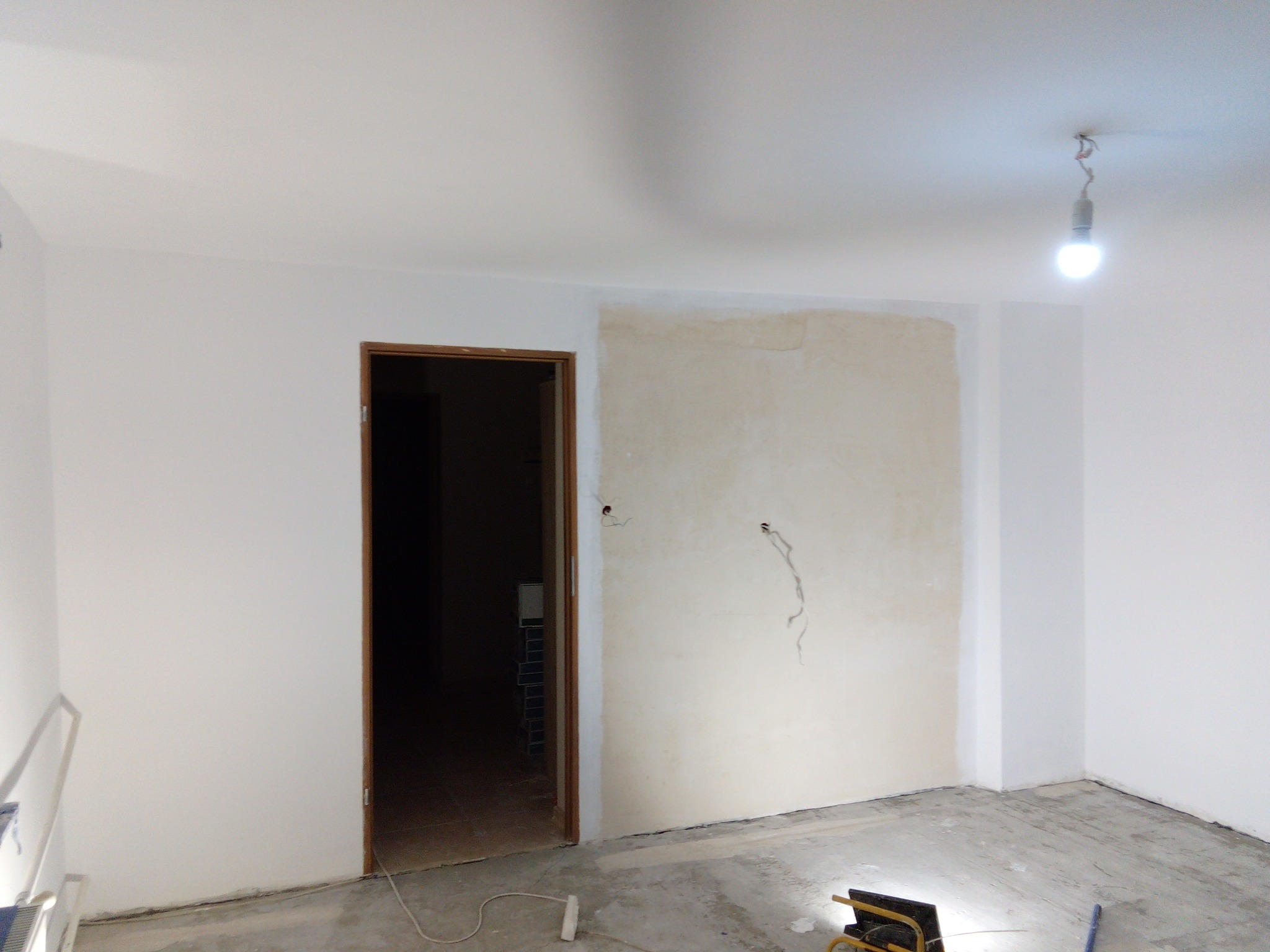 Pokój w trakcie odświeżania: surowe ściany z widocznymi przewodami elektrycznymi, częściowo nałożona warstwa wykończeniowa, goła żarówka zwisająca z sufitu, drewniane obramowanie drzwi, podłoga...