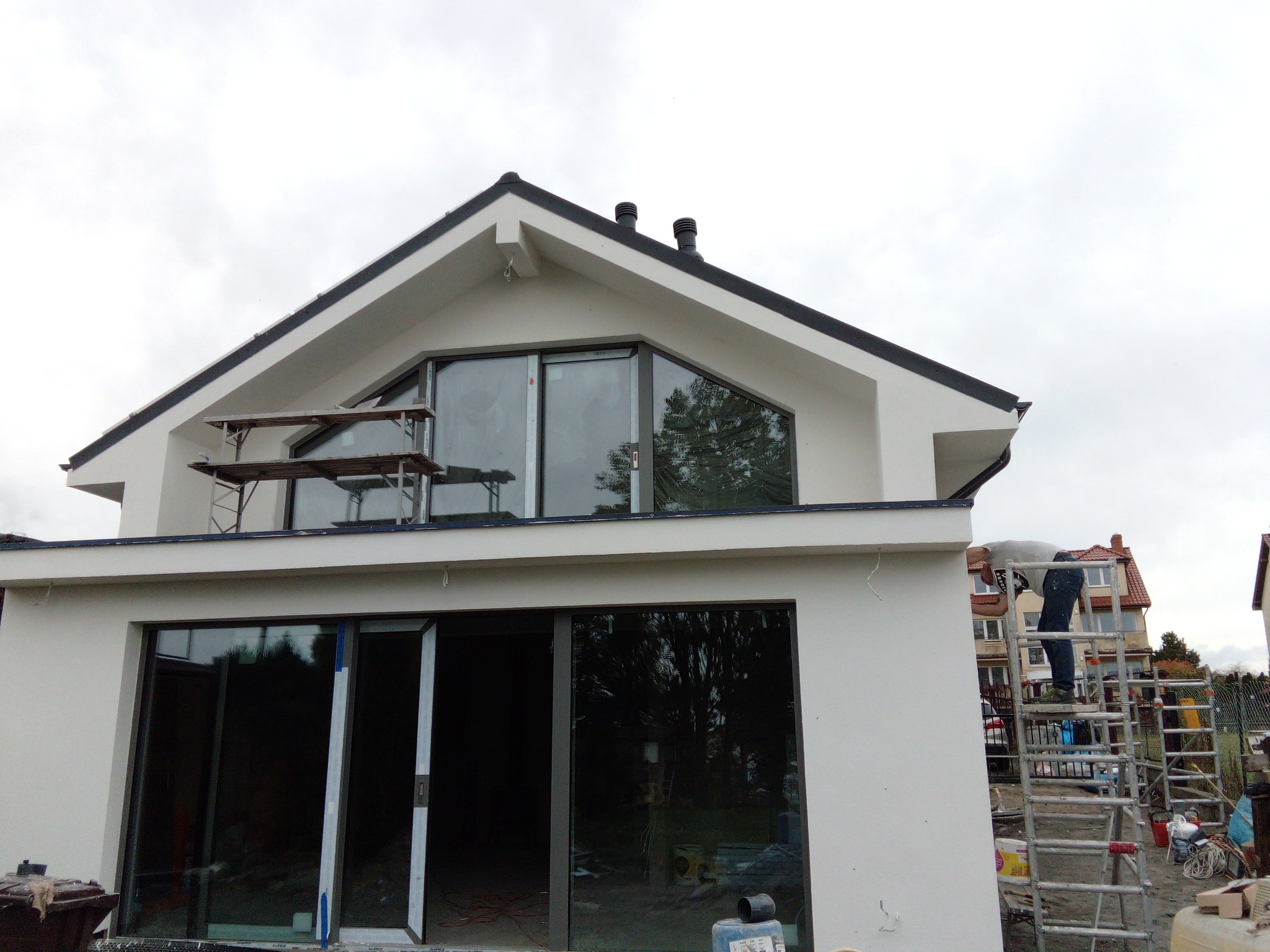 Nowoczesny dom w trakcie wykończenia elewacji, z widocznym rusztowaniem i pracownikiem na drabinie, duże okna od podłogi do sufitu, szare niebo w tle.