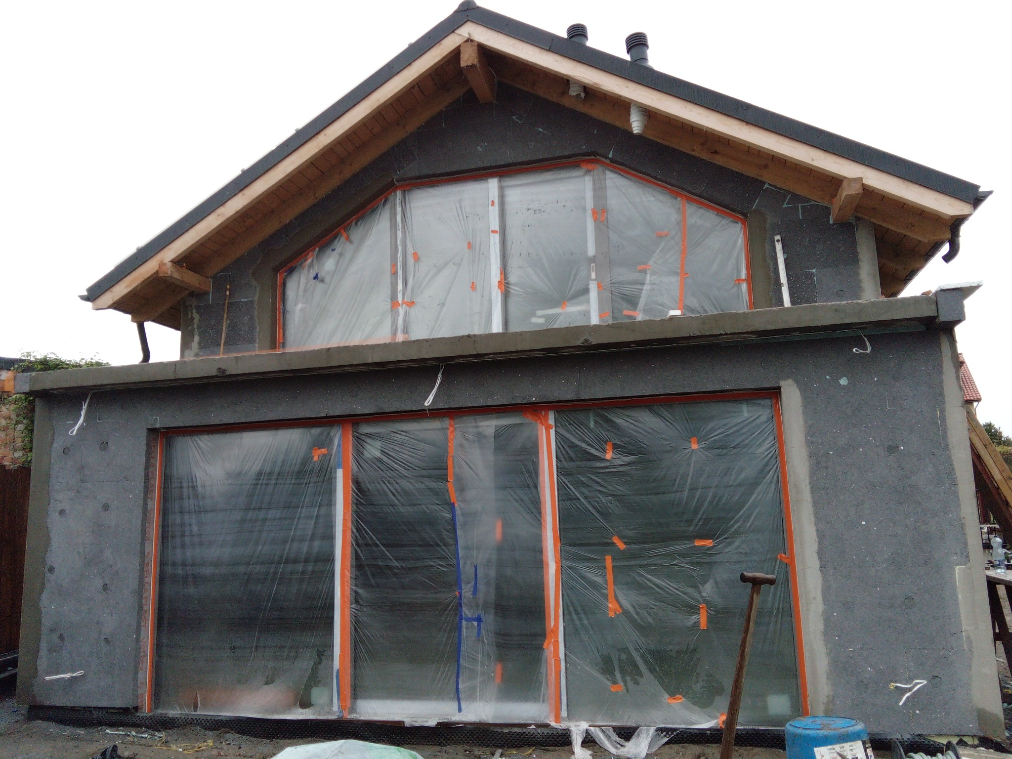 Surowa elewacja budynku w trakcie ocieplania, okna zabezpieczone folią, widoczne narzędzia i materiały budowlane.