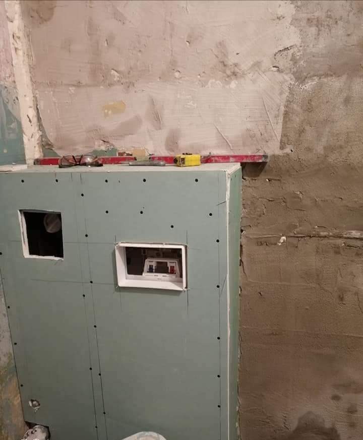 Montaż płyty karton gips z wyciętymi otworami na instalację elektryczną, widoczne narzędzia pracy: poziomica, miarka, okulary, surowa ściana w tle.