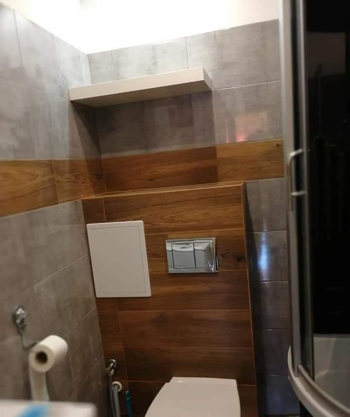 Wnętrze łazienki po remoncie: szare płytki ścienne, panel z płytek imitujących drewno, biała półka nad zabudową WC z chromowanym przyciskiem spłukiwania.