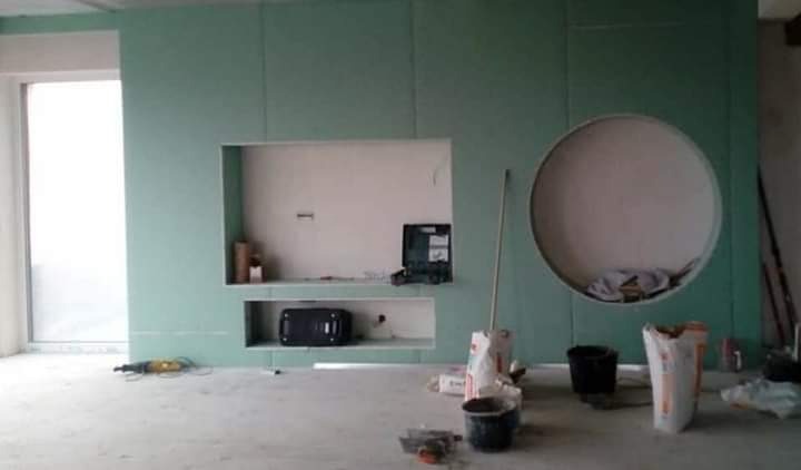 Ściana z płyty gipsowo-kartonowej w trakcie montażu, z widocznymi wnękami prostokątnymi i okrągłą, narzędzia i materiały budowlane na podłodze, surowe wykończenie.