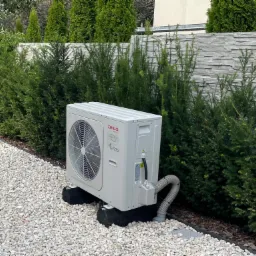 Biała jednostka zewnętrzna klimatyzacji Aiwa VR32 na czarnych gumowych podstawach, otoczona zielonymi krzewami i białym żwirem.