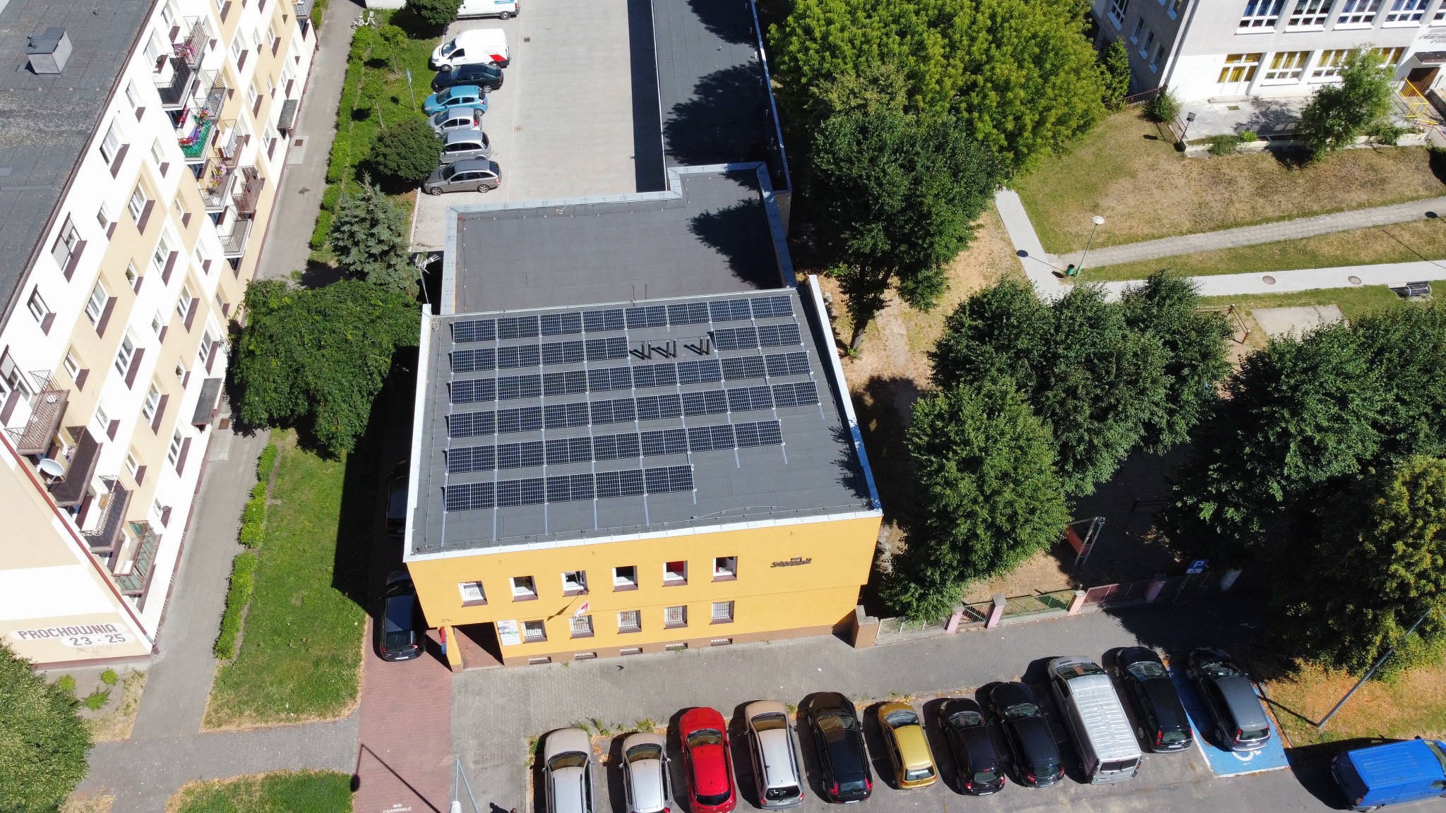 Widok z góry na budynek z żółtą elewacją i panelami fotowoltaicznymi na dachu, zaparkowane samochody wzdłuż budynku, zieleń i inne budynki w tle.