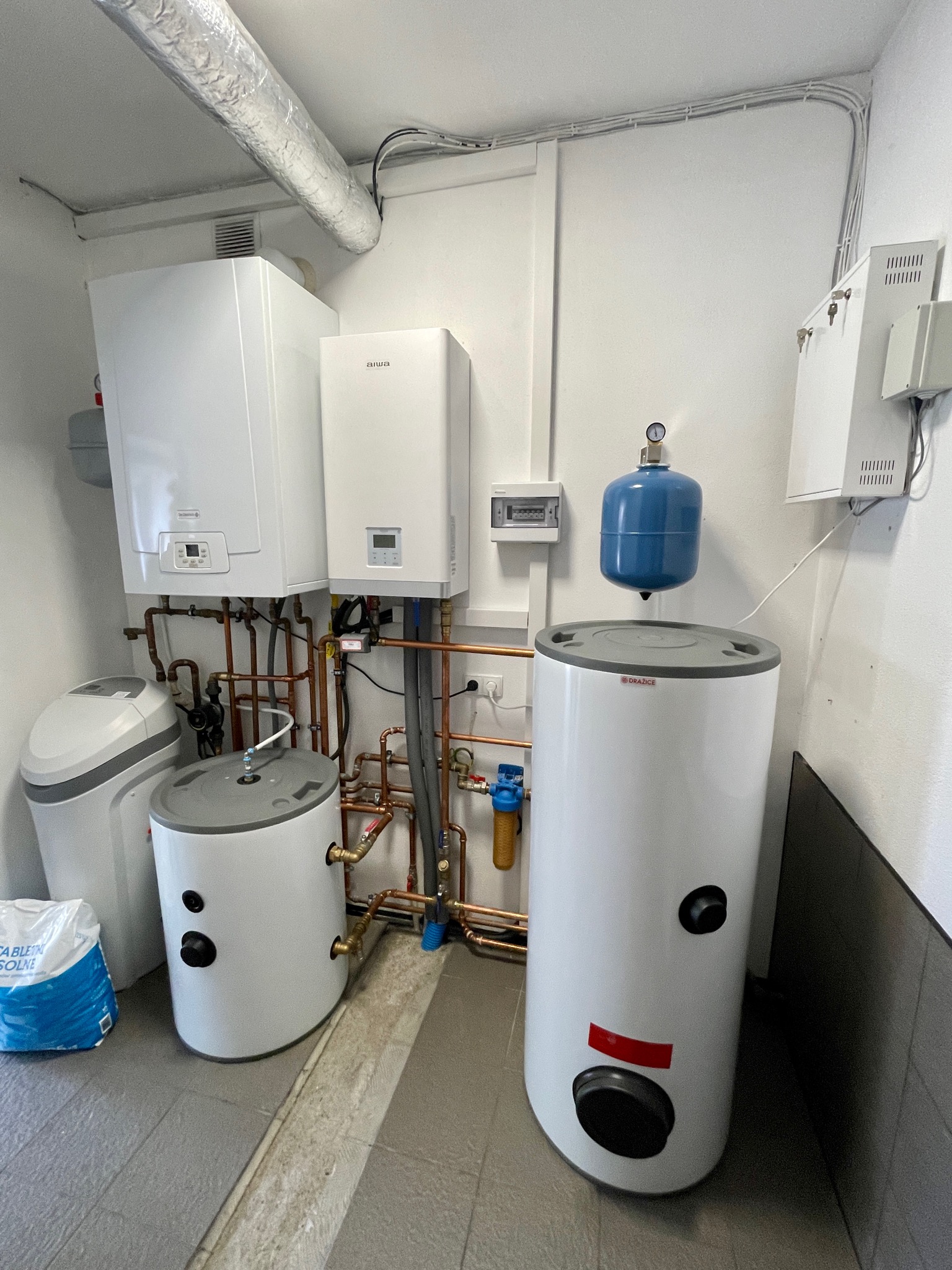 Kotłownia z dwoma białymi kotłami grzewczymi, dwoma zbiornikami na wodę, niebieskim naczyniem przeponowym, zmiękczaczem wody i workiem soli tabletkowej na szarej podłodze z płytek.