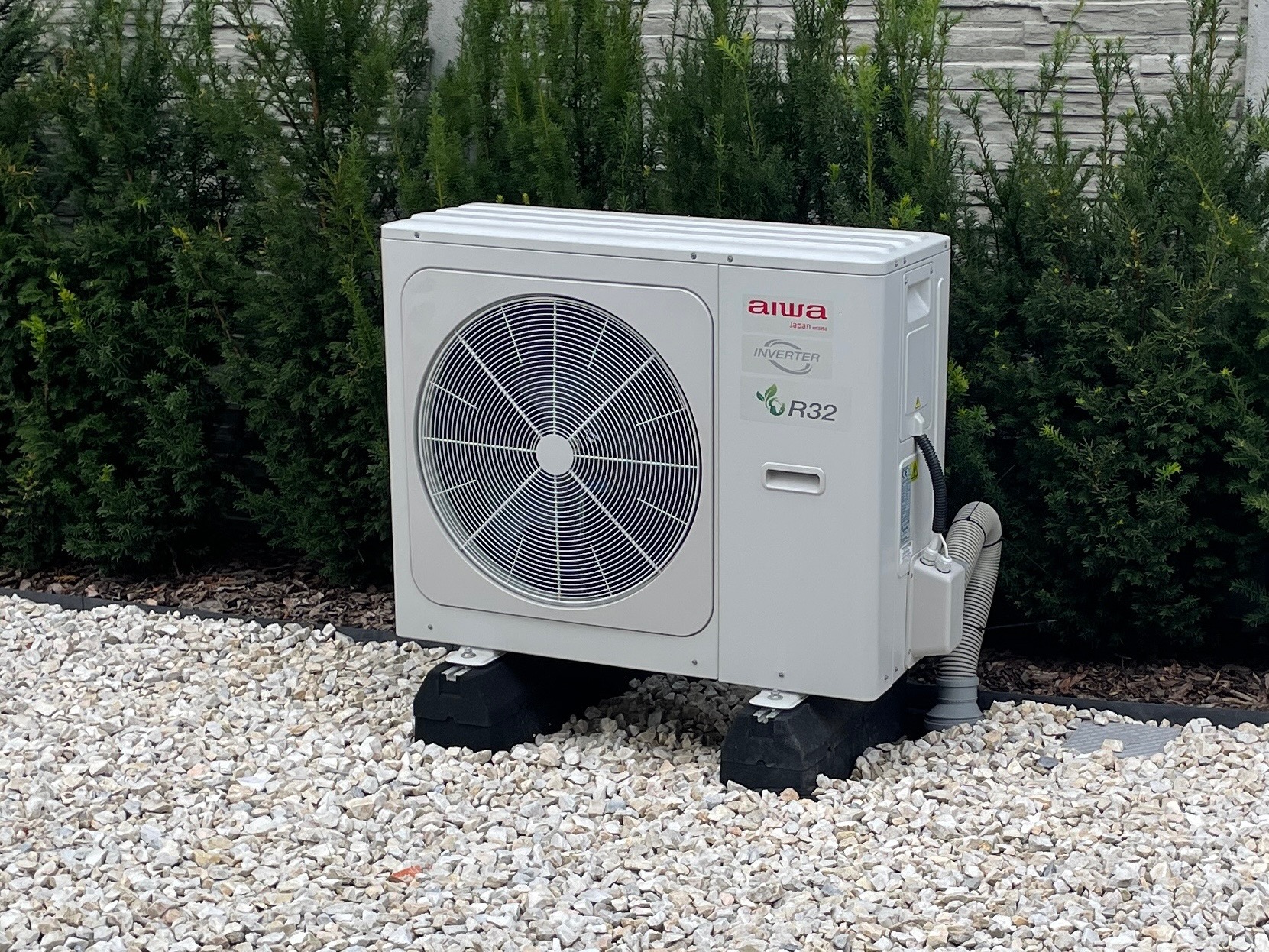 Biała jednostka zewnętrzna klimatyzacji Aiwa Inverter R32 na czarnych gumowych podstawach, otoczona białym żwirem i żywopłotem z tui.