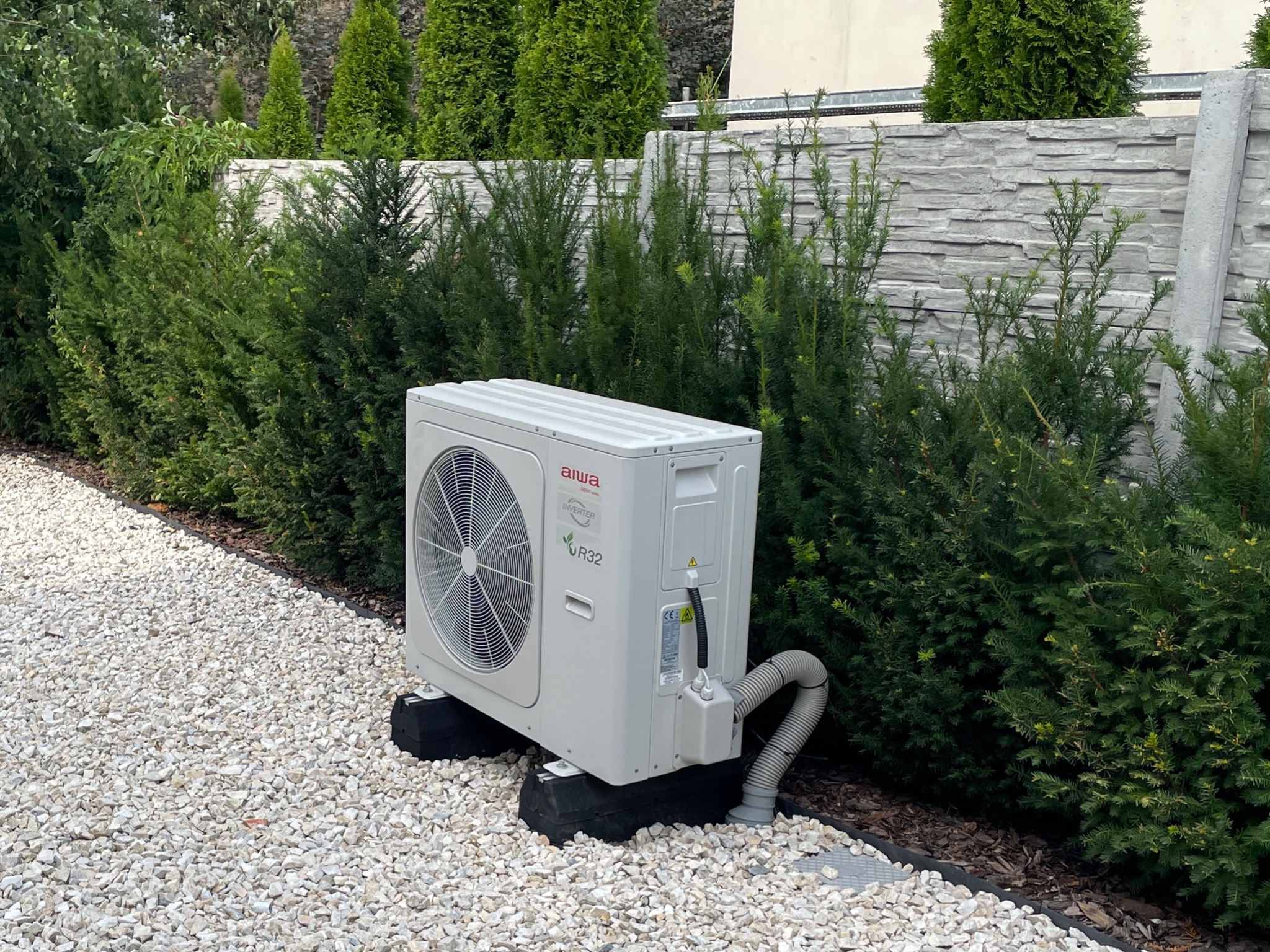 Biała jednostka zewnętrzna klimatyzacji Aiwa VR32 na czarnych gumowych podstawach, otoczona zielonymi krzewami i białym żwirem.