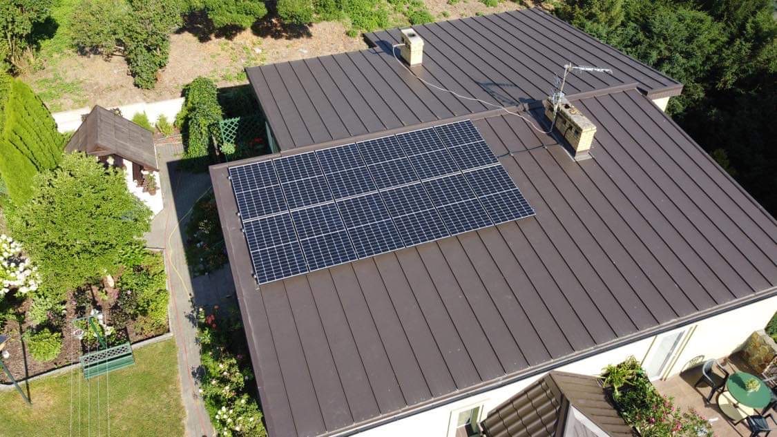 Widok z góry na brązowy dach z panelami słonecznymi, kominem i anteną, otoczony zielenią drzew i ogrodem.