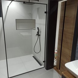 Barański Remonty i Wykończenia - Nowoczesna kabina prysznicowa walk-in z czarnymi elementami wykończeniowymi i odpływem liniowym. Białe płytki, prostokątna wnęka. Częściowo widoczna szafka z umywalką.