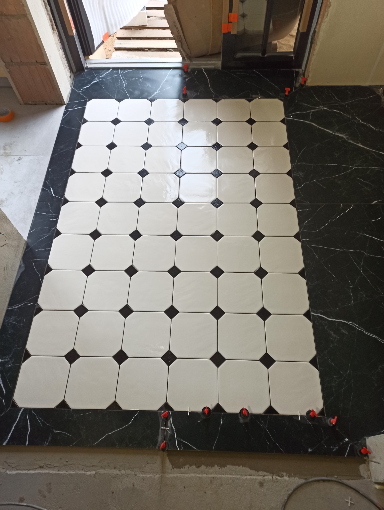 Układanie podłogi z płytek w biało-czarną szachownicę. Widoczne pomarańczowe kliny poziomujące. Czarny marmur na obrzeżach. Ujęcie z góry, w trakcie prac.