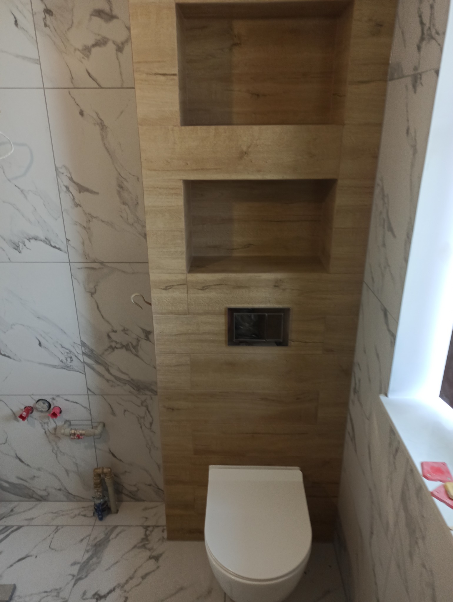 Wykończona łazienka z toaletą, drewnopodobnymi wnękami w ścianie i marmurowymi płytkami. Widoczne elementy instalacji wodnej. Minimalistyczny design.