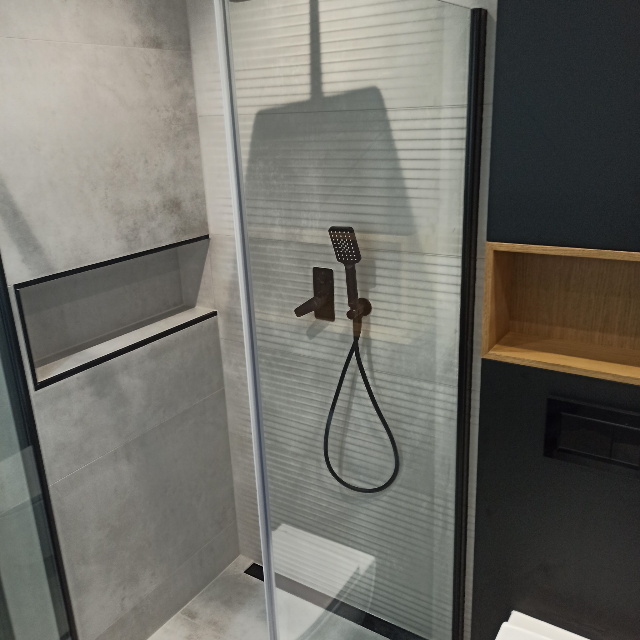 Nowoczesna kabina prysznicowa z deszczownicą w odcieniach szarości i czerni. Widoczna półka w ścianie i drewniana wnęka. Minimalistyczny design.
