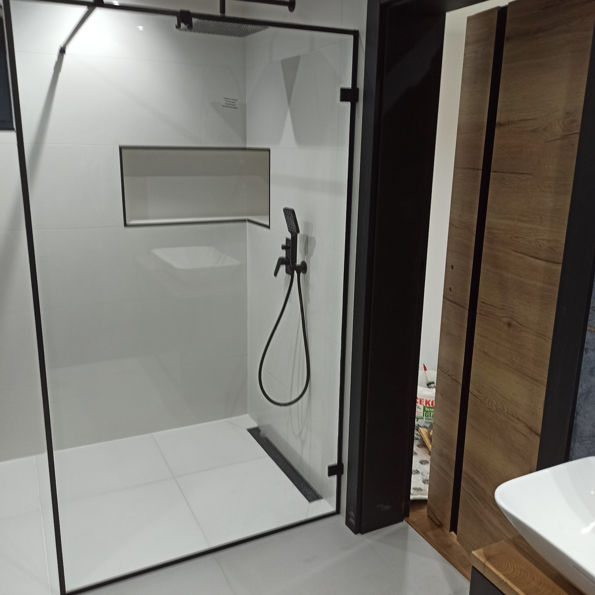 Nowoczesna kabina prysznicowa walk-in z czarnymi elementami wykończeniowymi i odpływem liniowym. Białe płytki, prostokątna wnęka. Częściowo widoczna szafka z umywalką.