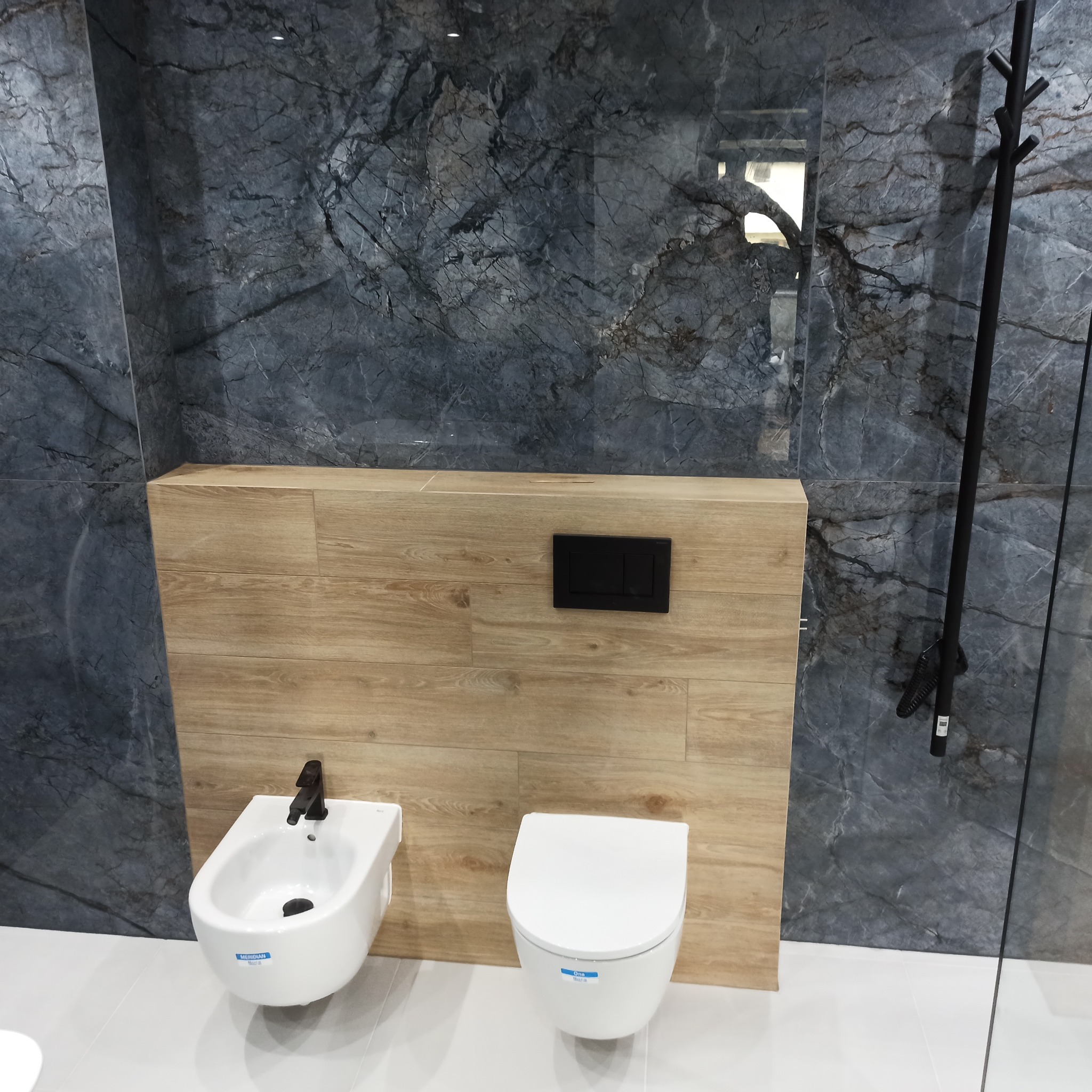 Nowoczesna łazienka z marmurowymi płytkami i drewnopodobną zabudową. Białe bidet i toaleta podwieszana z czarną armaturą. Minimalistyczny design.